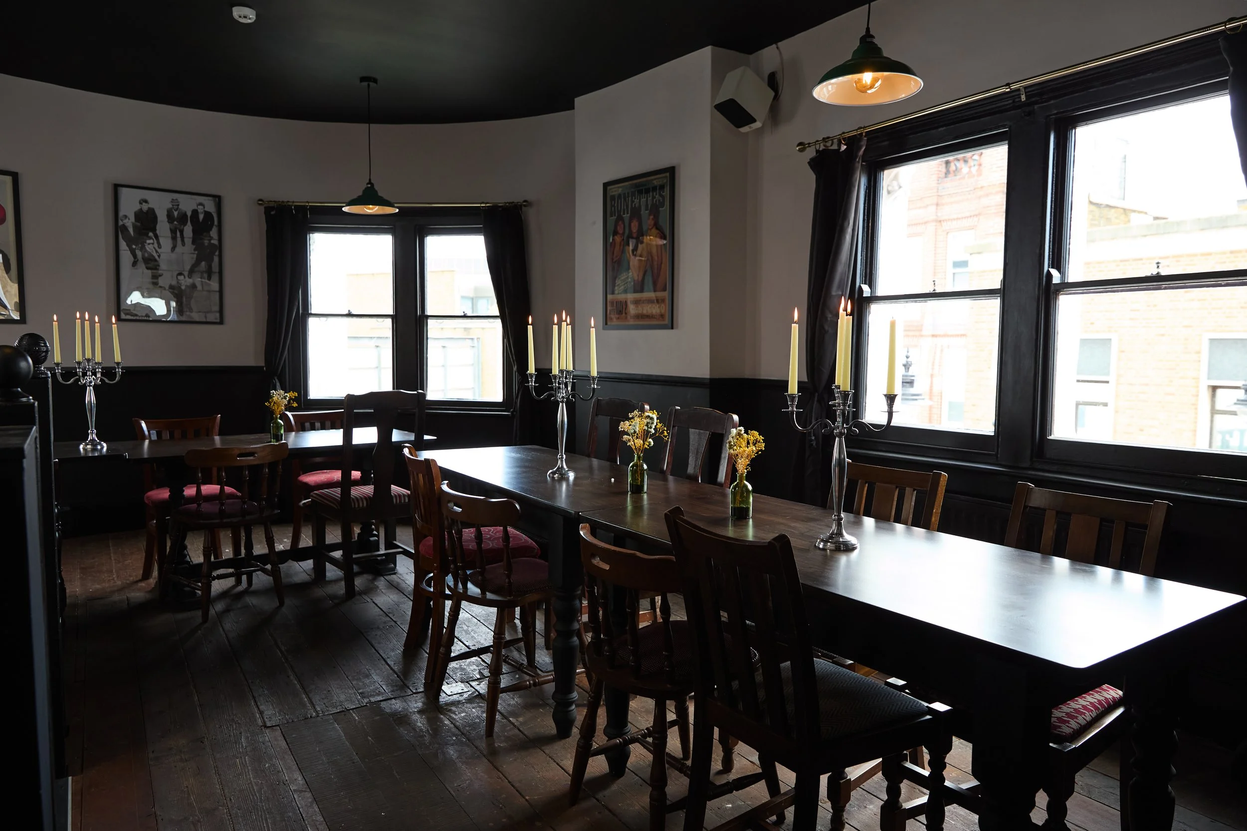 A cozy dining room with a large wooden table, multiple chairs, and candlesticks with lit candles, located near windows with dark curtains.