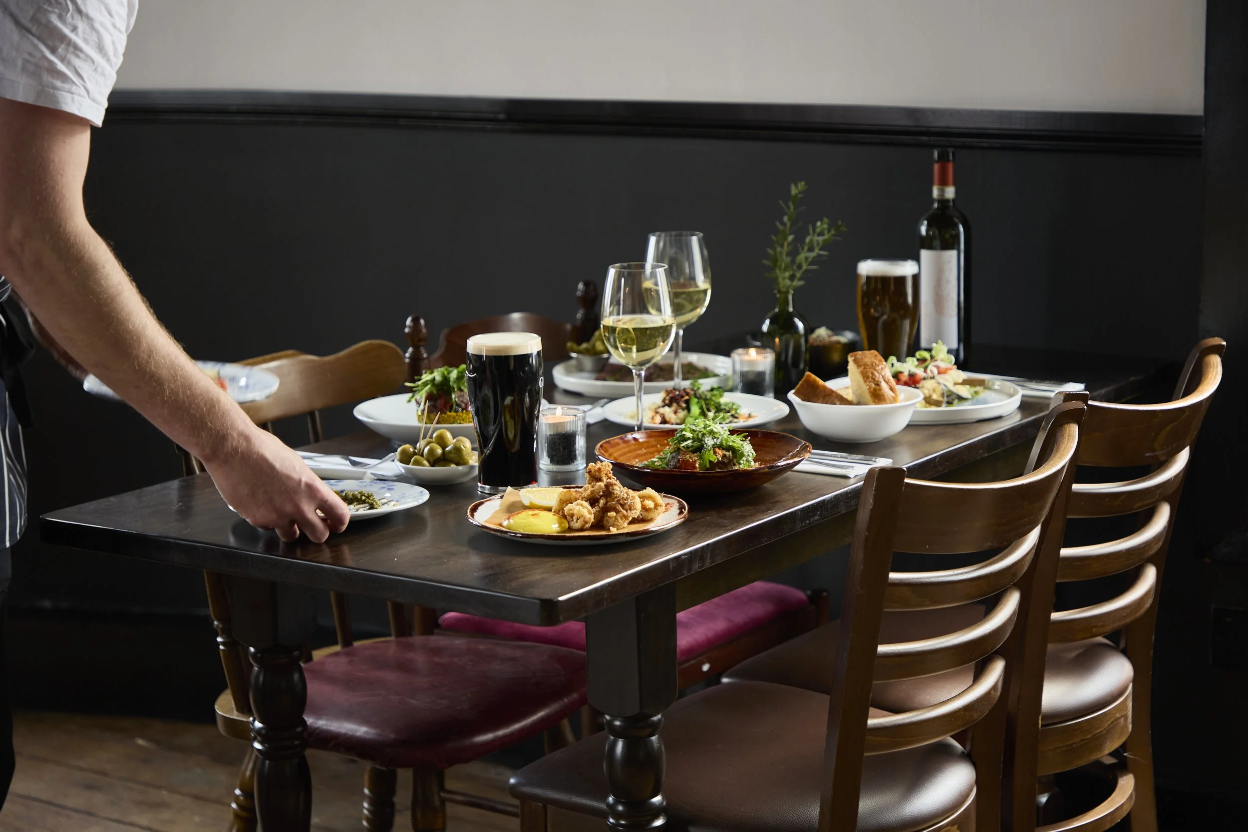 A restaurant table set with various dishes, drinks including wine, beer, and water, and some candles. There are chairs around the table and a person serving food.