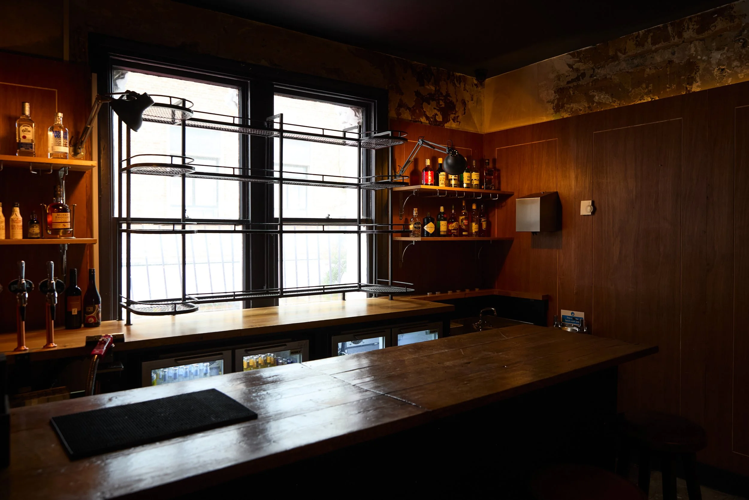 Empty bar with shelves of liquor bottles, wooden countertop, and large window with bars.