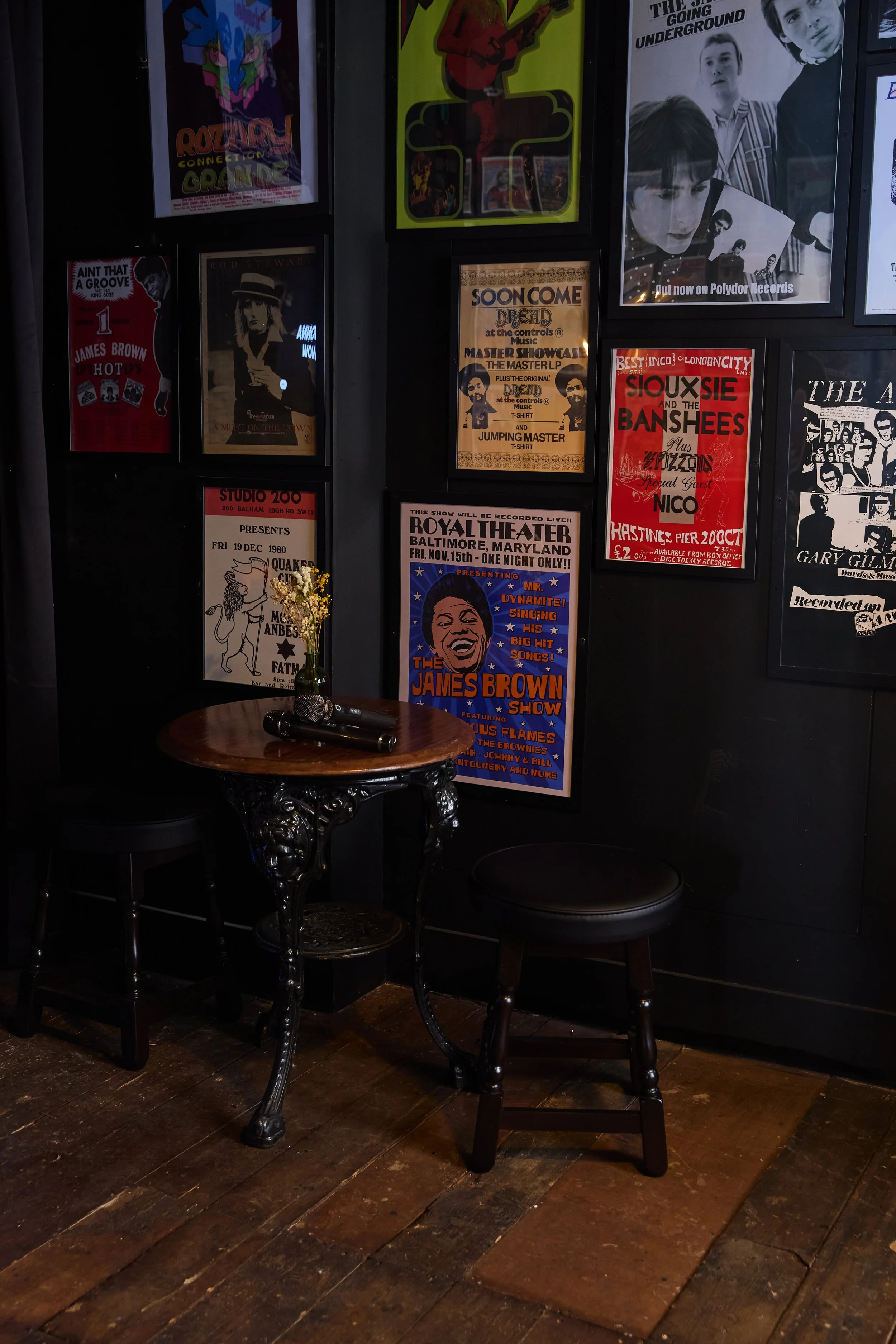Small round table with a small vase of yellow flowers and two microphones, surrounded by two black chairs, against a black wall adorned with vintage concert and event posters.