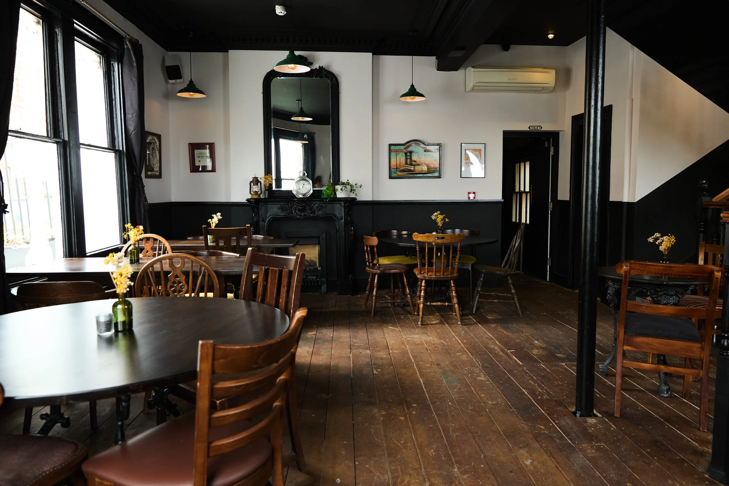 Interior of a cozy cafe with wooden floors, several round and square tables, each decorated with small vases of flowers, black and white walls, and vintage framed art. Large windows with black trim allow natural light to fill the space. There is a black ornate fireplace mantel with decorative items and a mirror above it. Pendant lights hang from the ceiling, and an air conditioning unit is visible on the wall.