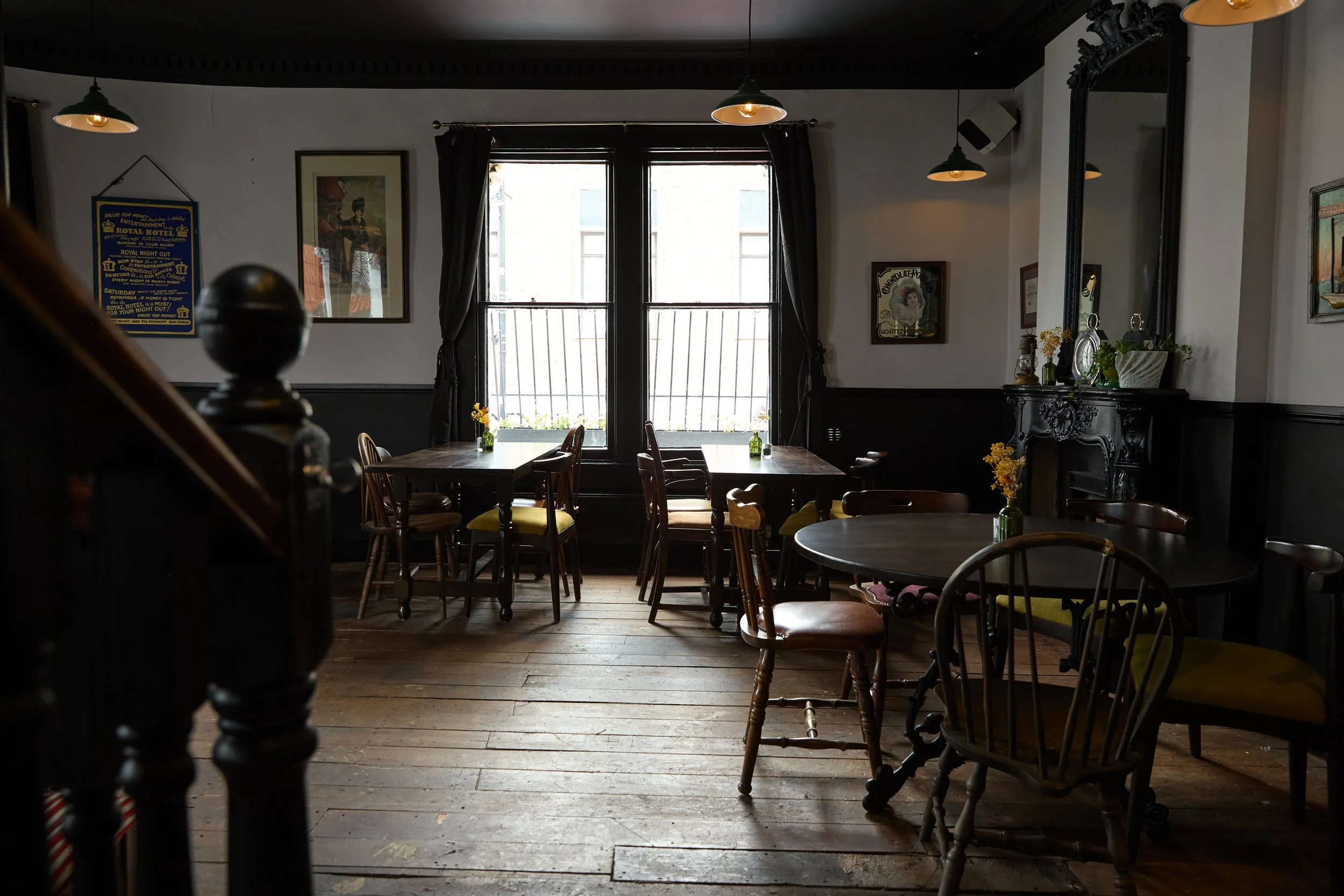 Interior of a cozy cafe or restaurant with wooden floors, dark wainscoting, and vintage decor. There are several tables with wooden chairs, some with yellow cushions, and small vases with yellow flowers. Large windows with dark curtains allow natural