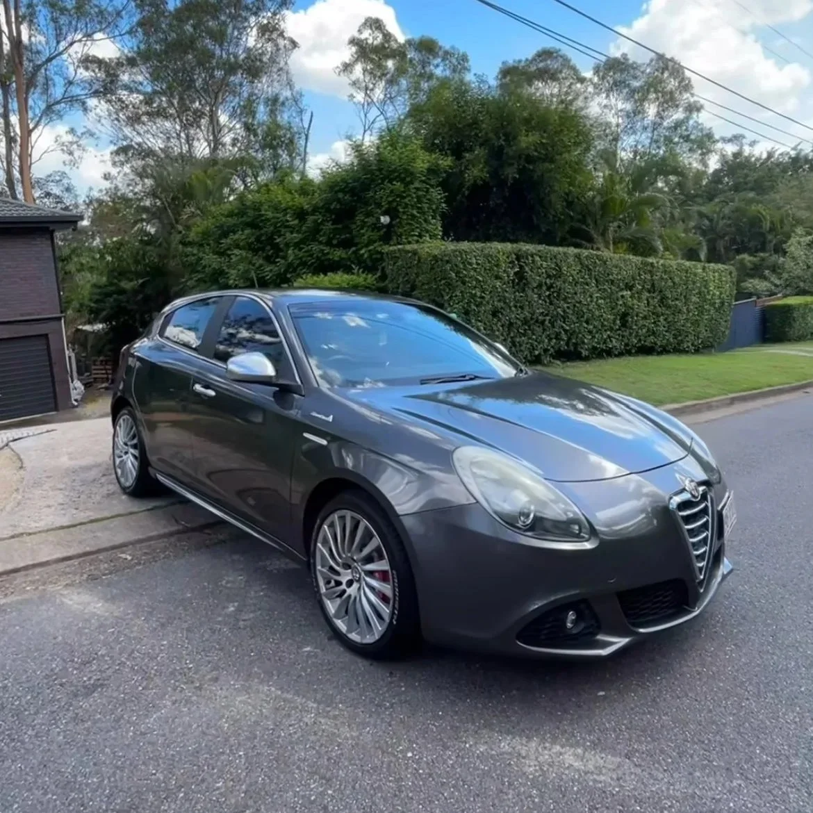 A dark gray Alfa Romeo hatchback car parked on a residential street with green bushes and trees in the background.