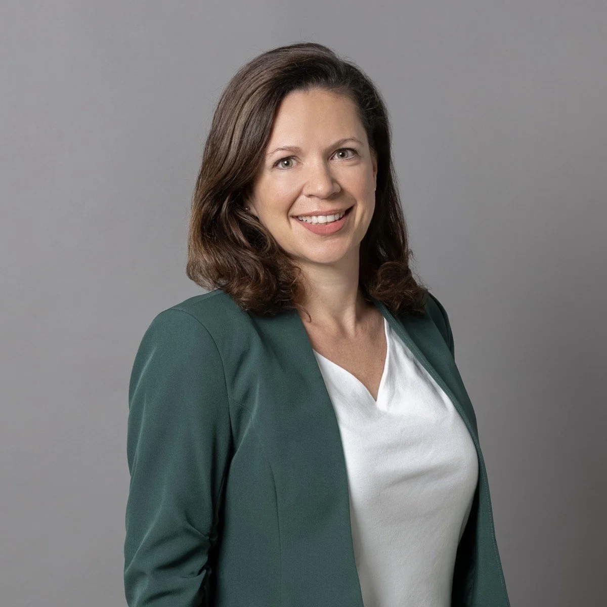 Porträt einer lächelnden Frau mit braunen, gewellten Haaren, trägt eine weiße Bluse und eine dunkelgrüne Jacke, vor einem grauen Hintergrund.