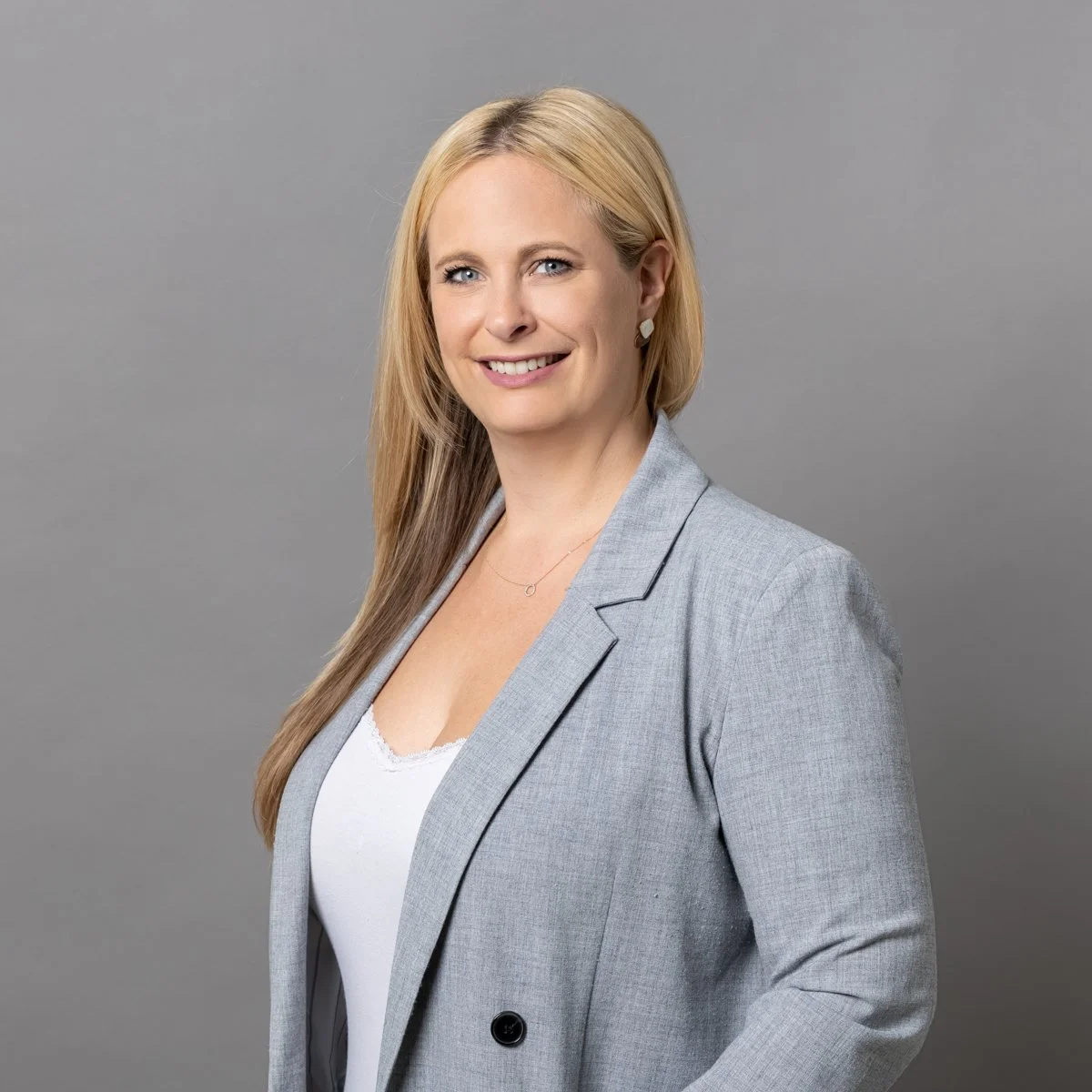Porträt einer lächelnden Frau mit blonden Haaren in grau blazer vor grauem Hintergrund.