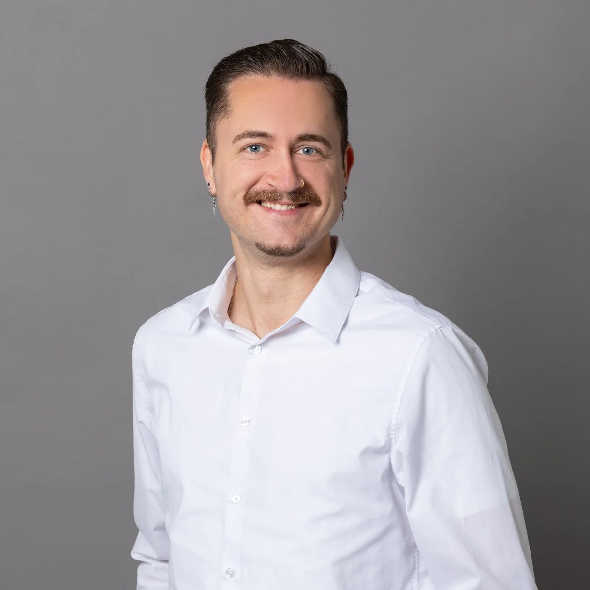 Ein lachender Mann mit dunklem, glattem Haar, Schnurrbart und Piercings, trägt ein weißes Hemd vor einem grauen Hintergrund.