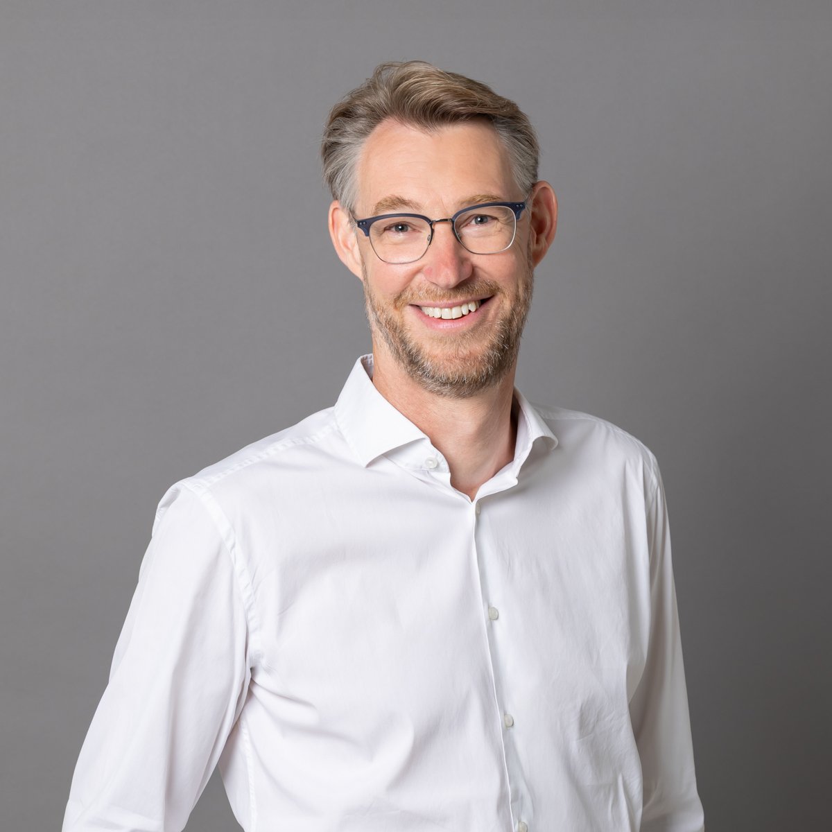 Ein Mann mit Brille, grauem Haar und Bart trägt ein weißes Hemd und lächelt vor grauem Hintergrund.