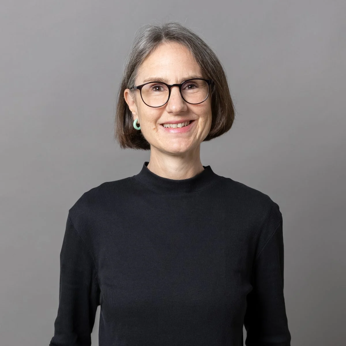 Porträt einer lächelnden Frau mit schwarzen Brillen, kurzen braunen Haaren, weißen Zähnen, einem schwarzen Rollkragenpullover, einer grünen Ohrring und einem neutralgrauen Hintergrund.