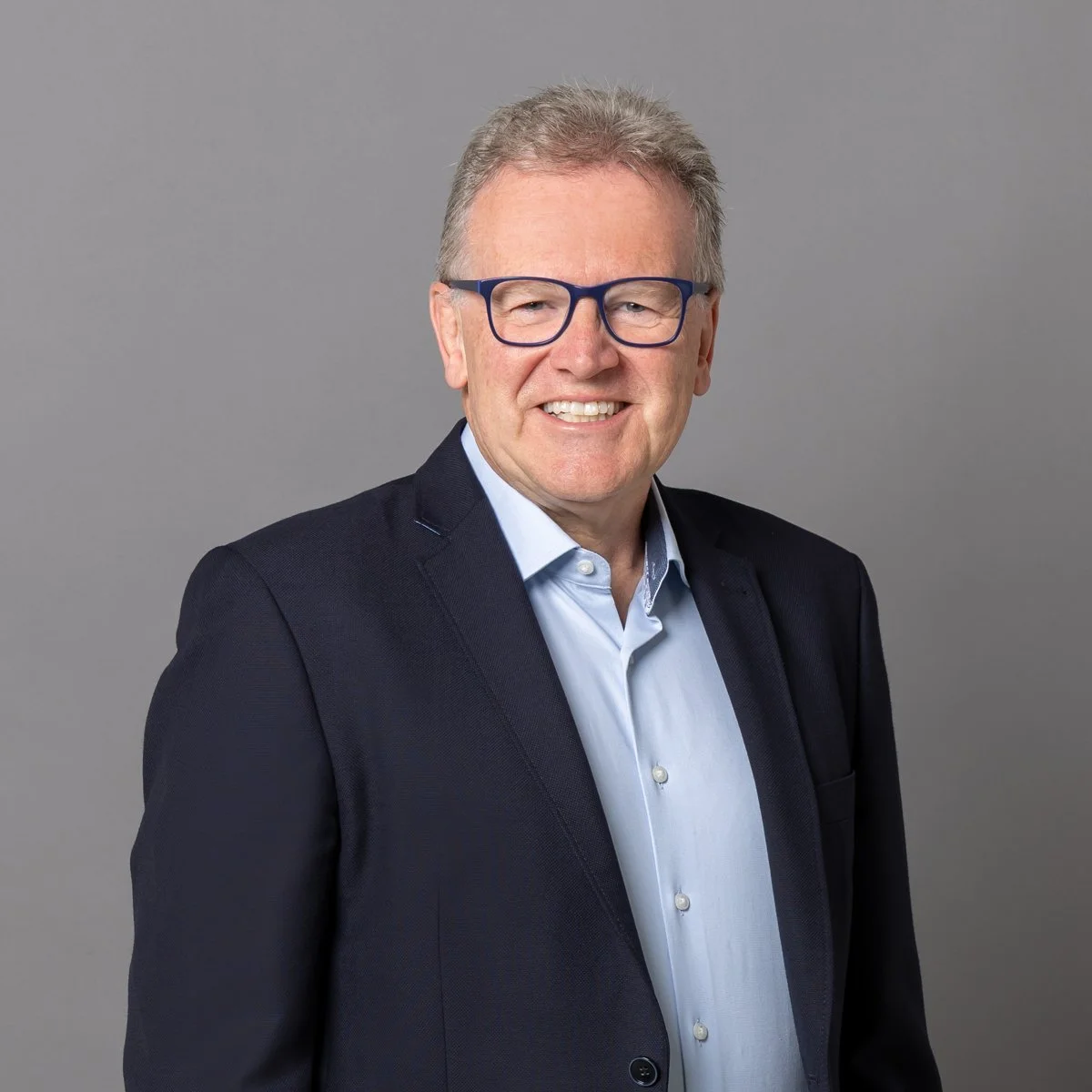 Ein Mann in formeller Kleidung mit Brille vor grauem Hintergrund, lächelnd.