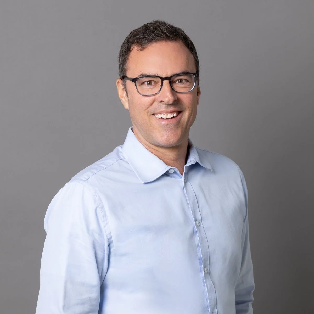 Ein Mann mit dunklem, kurzem Haar, Brille und freundlichem Lächeln trägt ein hellblaues Hemd vor einem grauen Hintergrund.