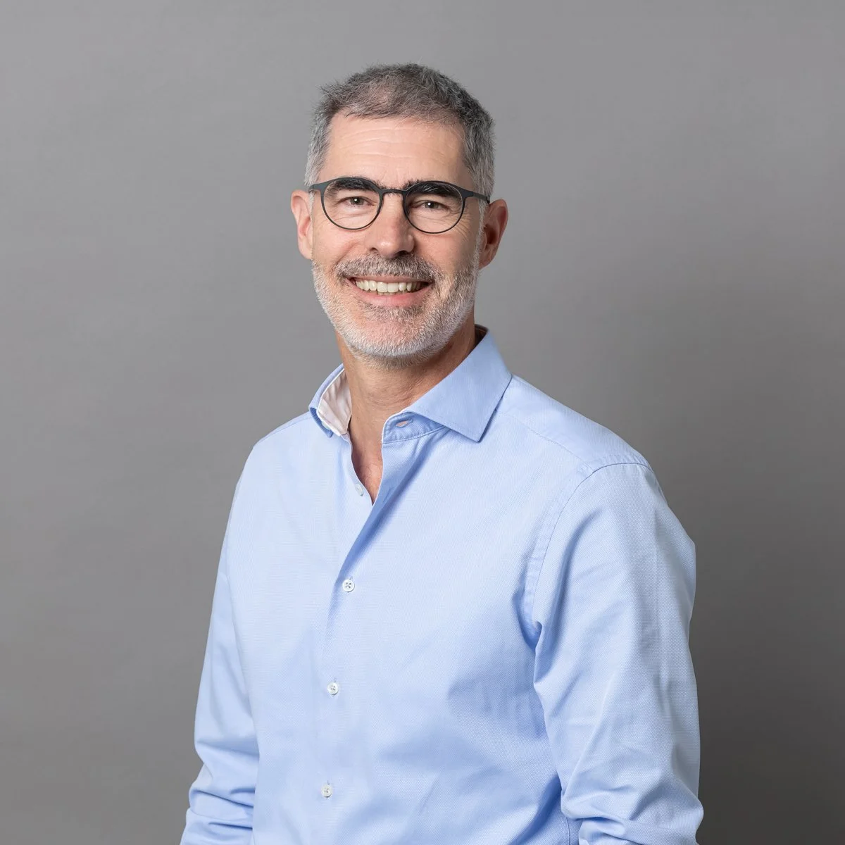 Ein lächelnder Mann mit Brille und grauem Haar trägt ein hellblaues Hemd vor einem grauen Hintergrund.