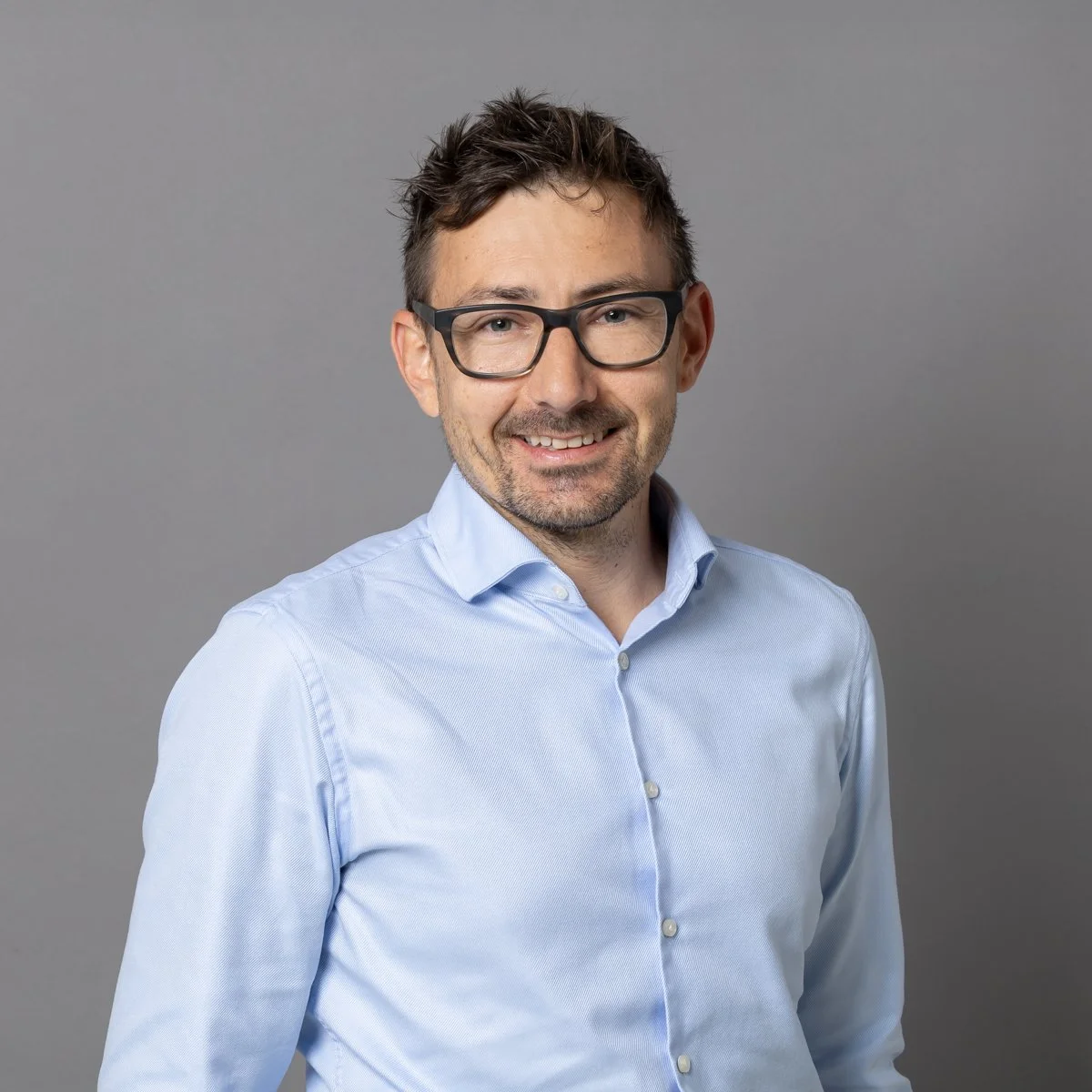 Ein Mann mit Brille in einem blauen Hemd vor grauem Hintergrund.