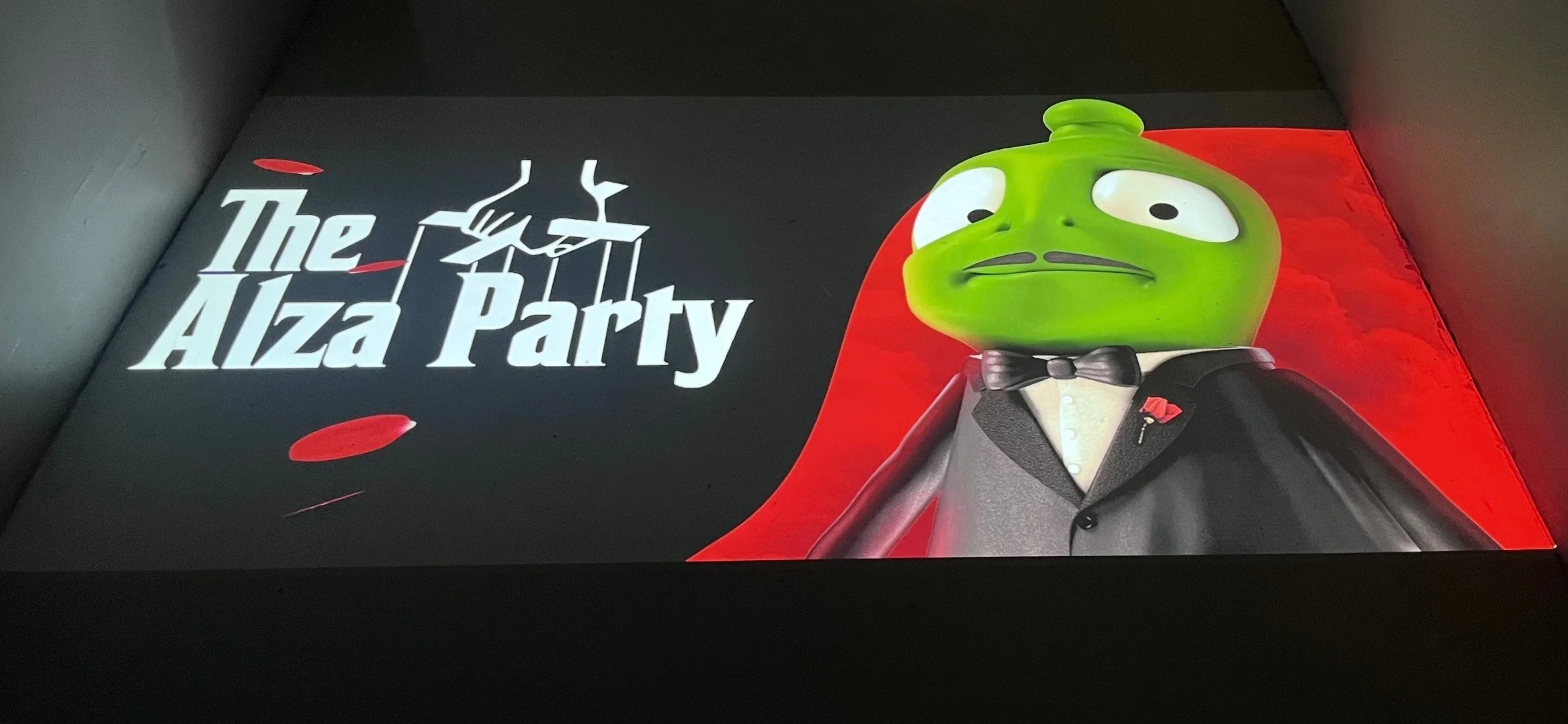 Egy zöld bábura hasonlító, öltönyös, csokornyakkendős figura vetítése az „Alza Party” felirat mellett, bábkéz motívummal, ami a keresztapa stílusát idézi.