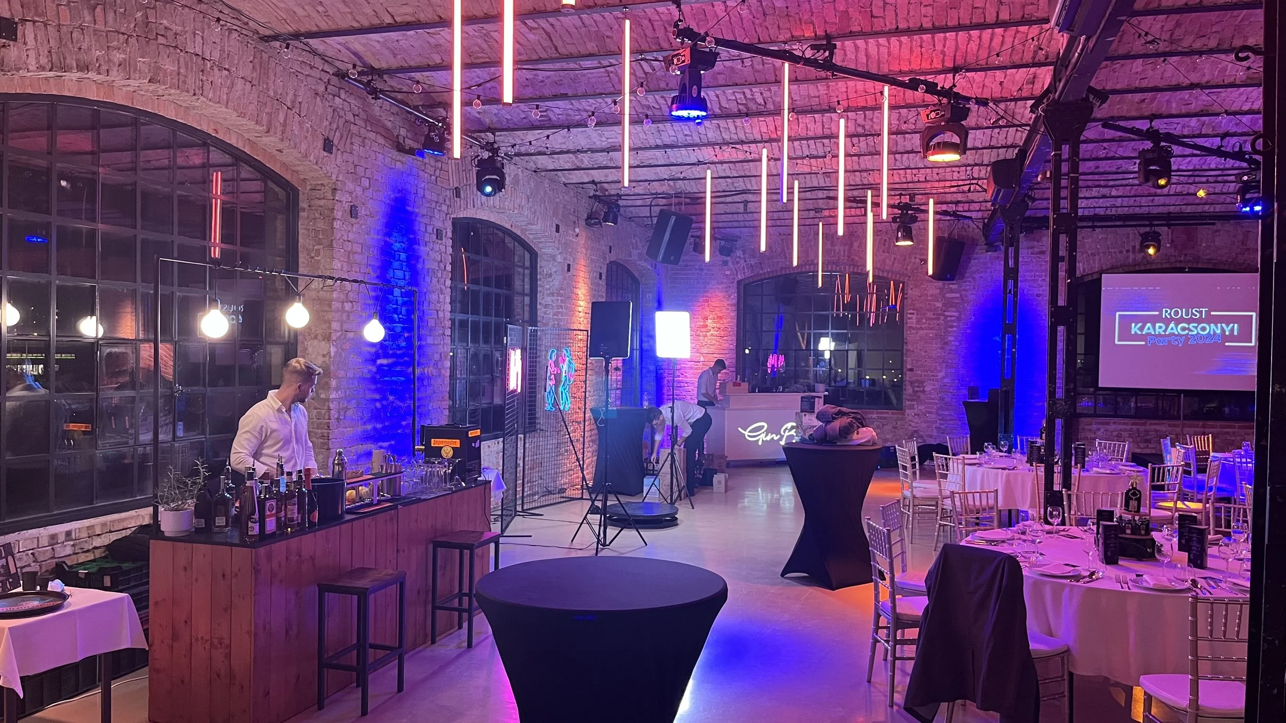 Rendezvényhelyszín asztalokkal és bárral egy ipari stílusú helyszínen, ahol látható téglafalak, lógó fények és egy vetítővászon. Az asztalok meg vannak terítve az étkezéshez, és egy pultos készíti az italokat.