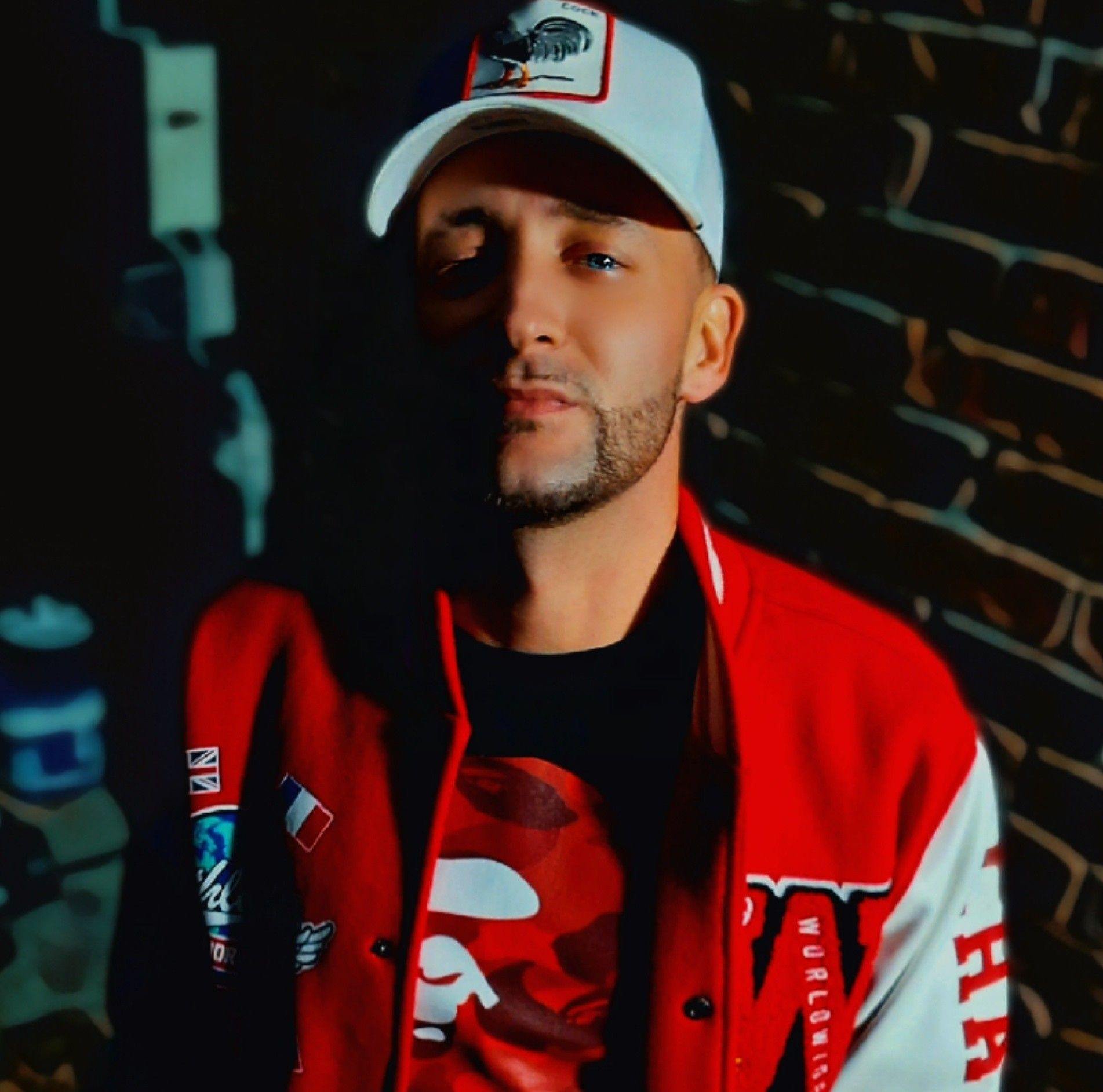 Portrait of Louisiana underground hip-hop artist Chevy Jade standing against a dark brick wall. He is wearing a white graphic baseball cap, a red and white varsity jacket with patches, and a red camo t-shirt under dramatic lighting.