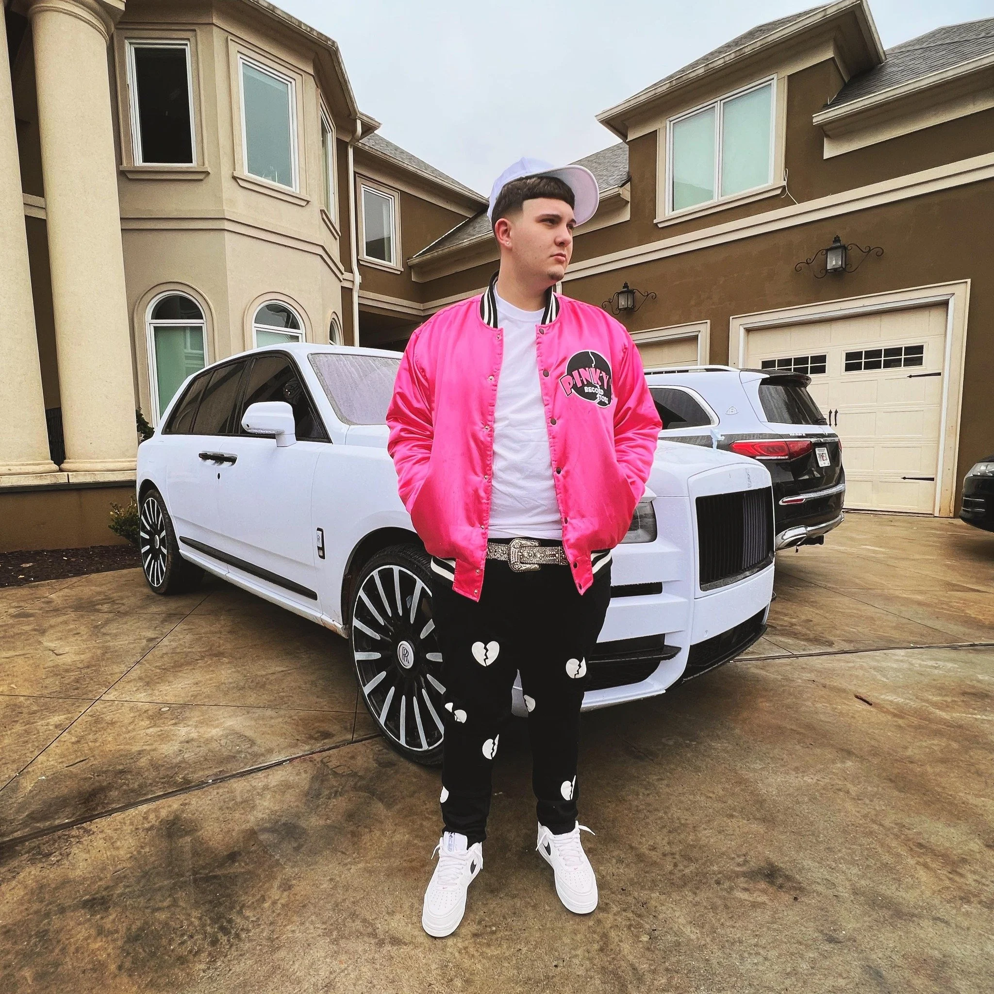 LD Visuals, premier Baton Rouge videographer, standing in front of a white Rolls-Royce Cullinan outside a luxury estate. He is wearing a pink bomber jacket and custom black pants.