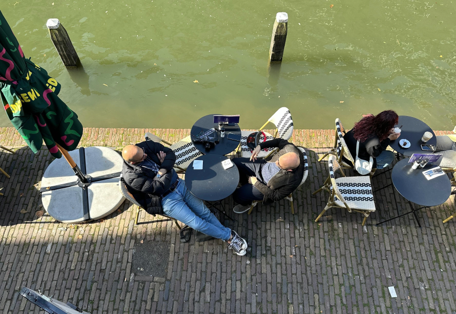 People sitting at outdoor tables by a canal, with a large umbrella, some empty chairs, and people talking and eating.