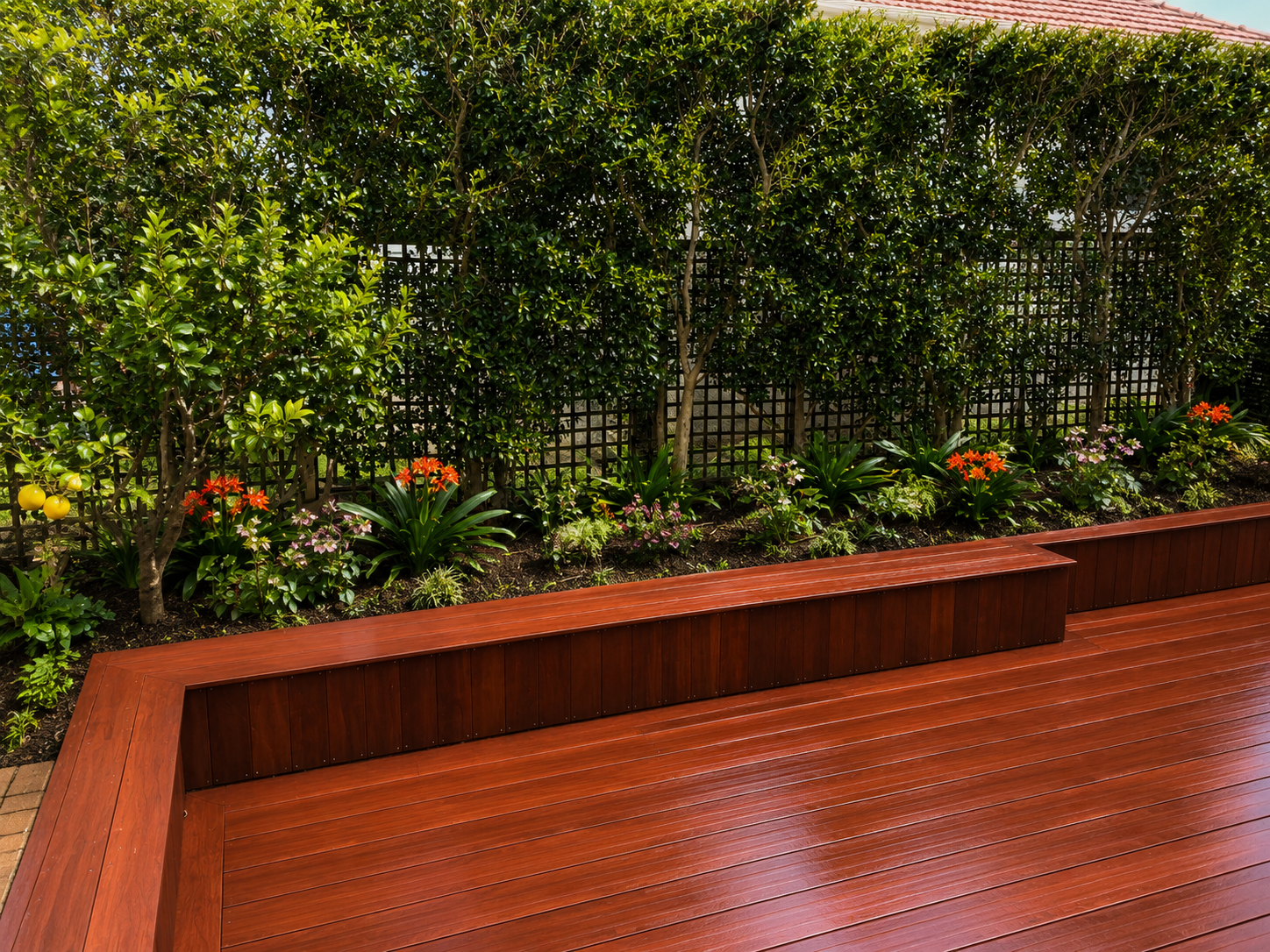 Wooden deck with built-in bench, a landscaped garden with various green plants, bushes, and colorful flowers, and a tall hedge with lattice behind it in an outdoor setting.