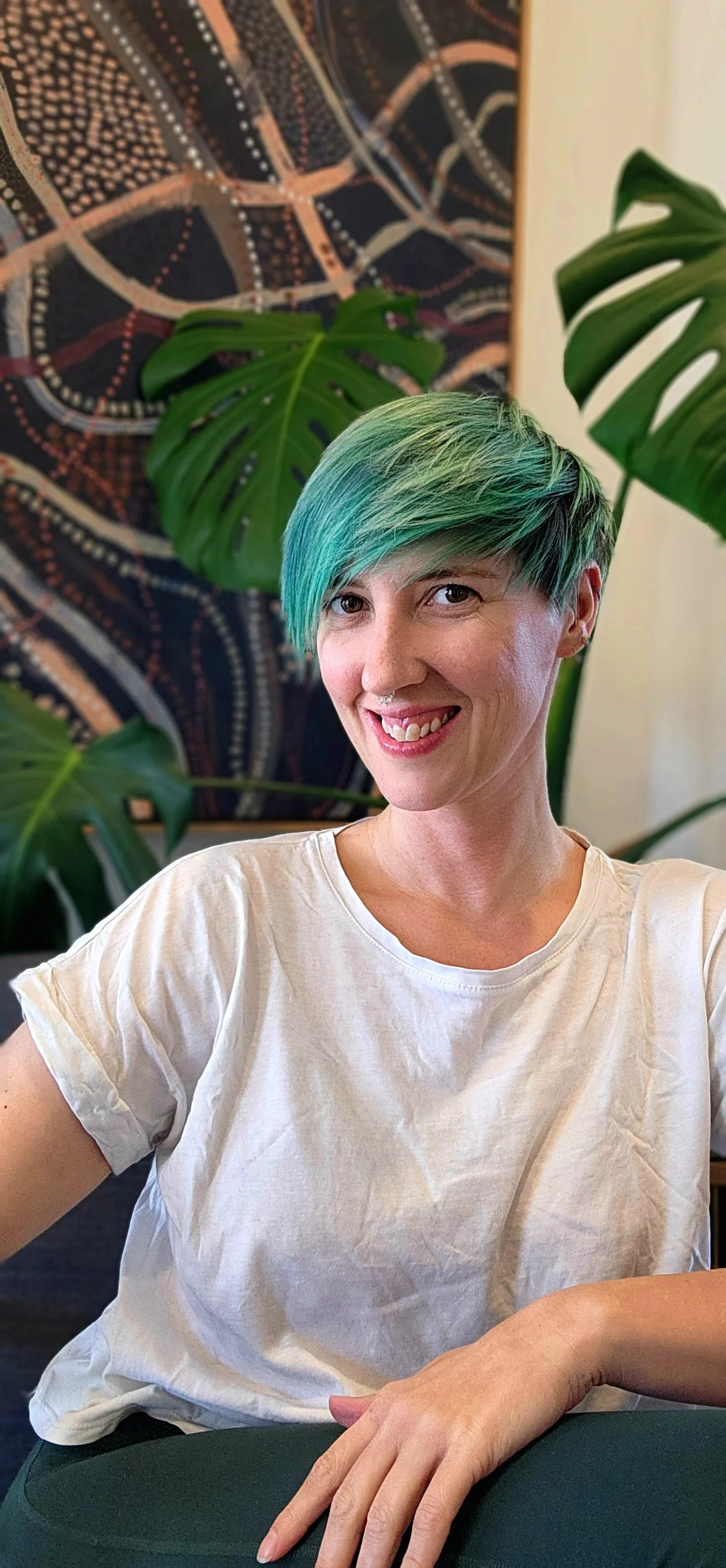 Smiling woman with short, turquoise hair sitting in front of green plants and an abstract artwork.
