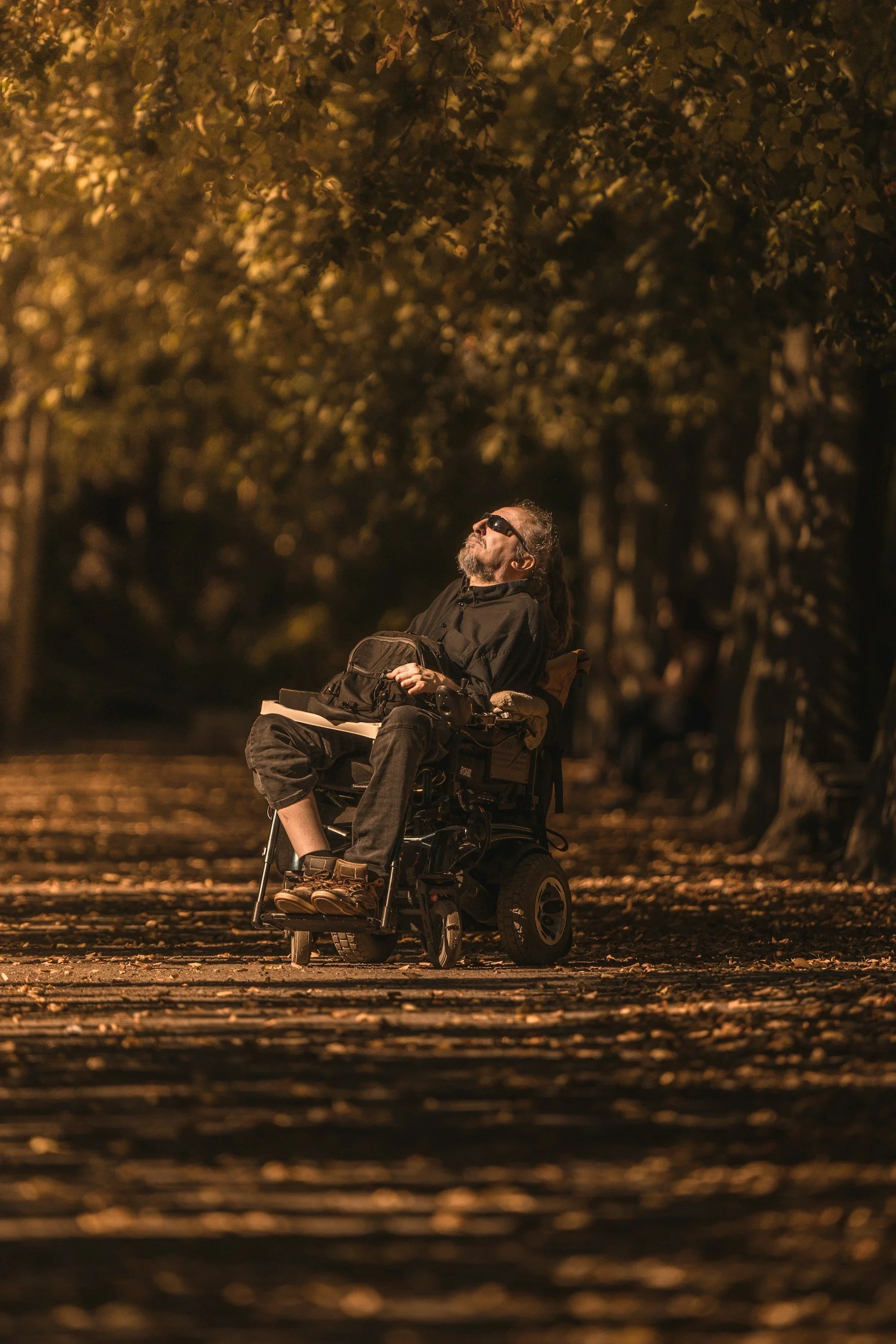 A man with long gray hair and a beard sitting in a motorized wheelchair on a path surrounded by trees with golden leaves, wearing sunglasses and a black outfit, looking up at the sky.