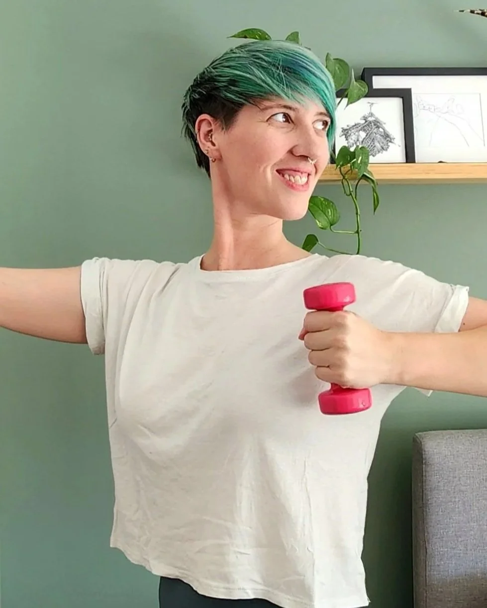 A woman with short, teal hair holding a pink dumbbell and smiling while flexing her arm.