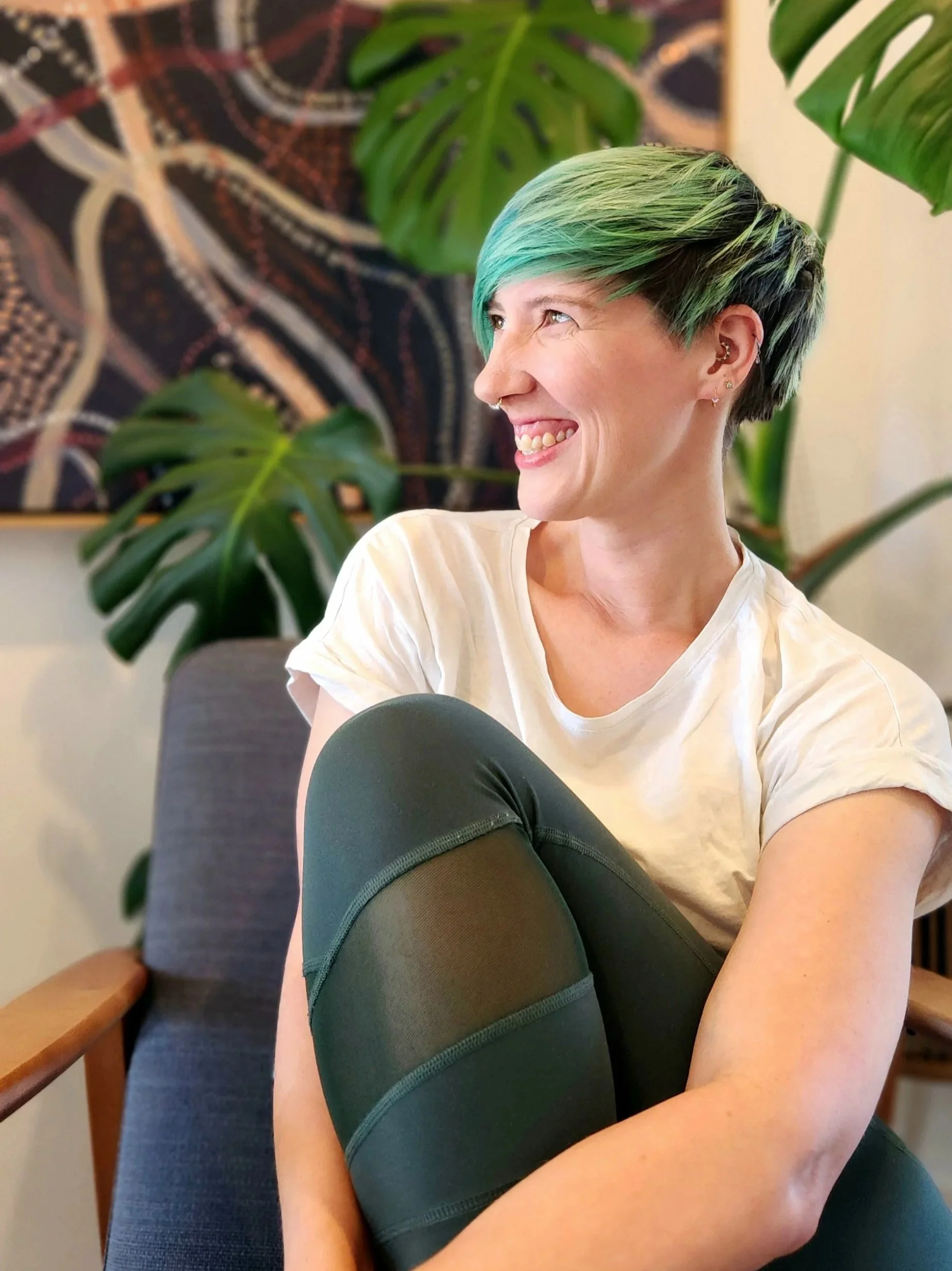 A woman with short green hair, wearing a white t-shirt and green leggings, smiles while sitting in a grey chair inside in front of a large painting with greenery.