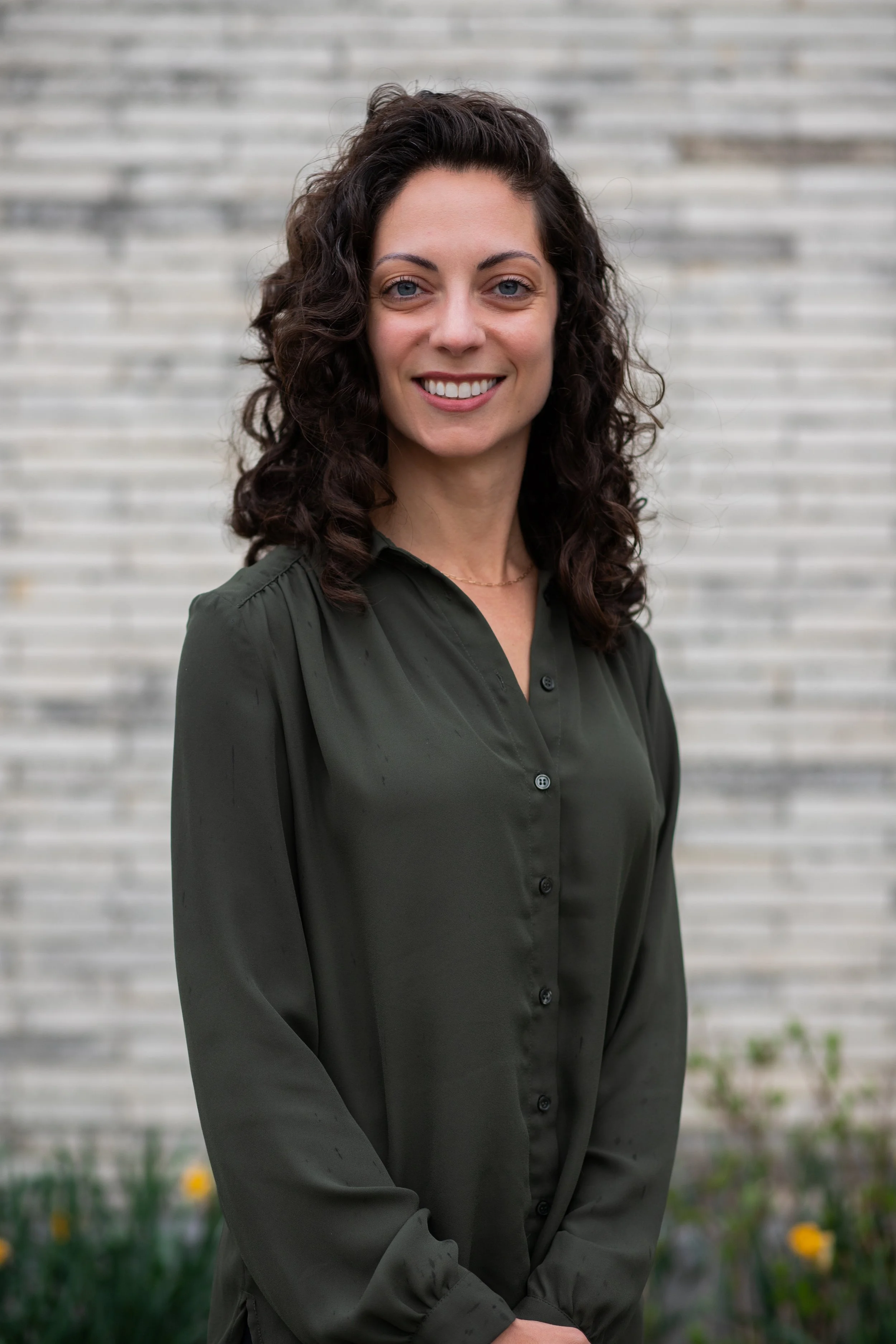 A woman with curly brown hair and light skin, smiling, standing outdoors in front of a light-colored brick wall. She is wearing a dark green button-up blouse.