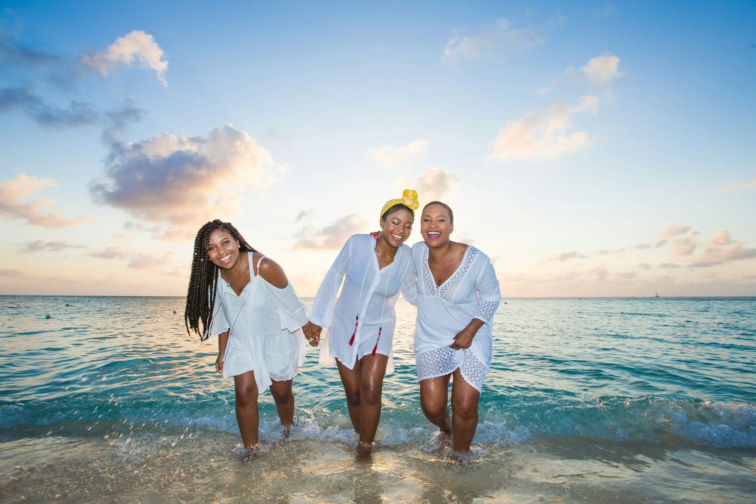 Three Black women in white dresses holding hands and walking in shallow ocean water at sunset, smiling and enjoying a beach outing.