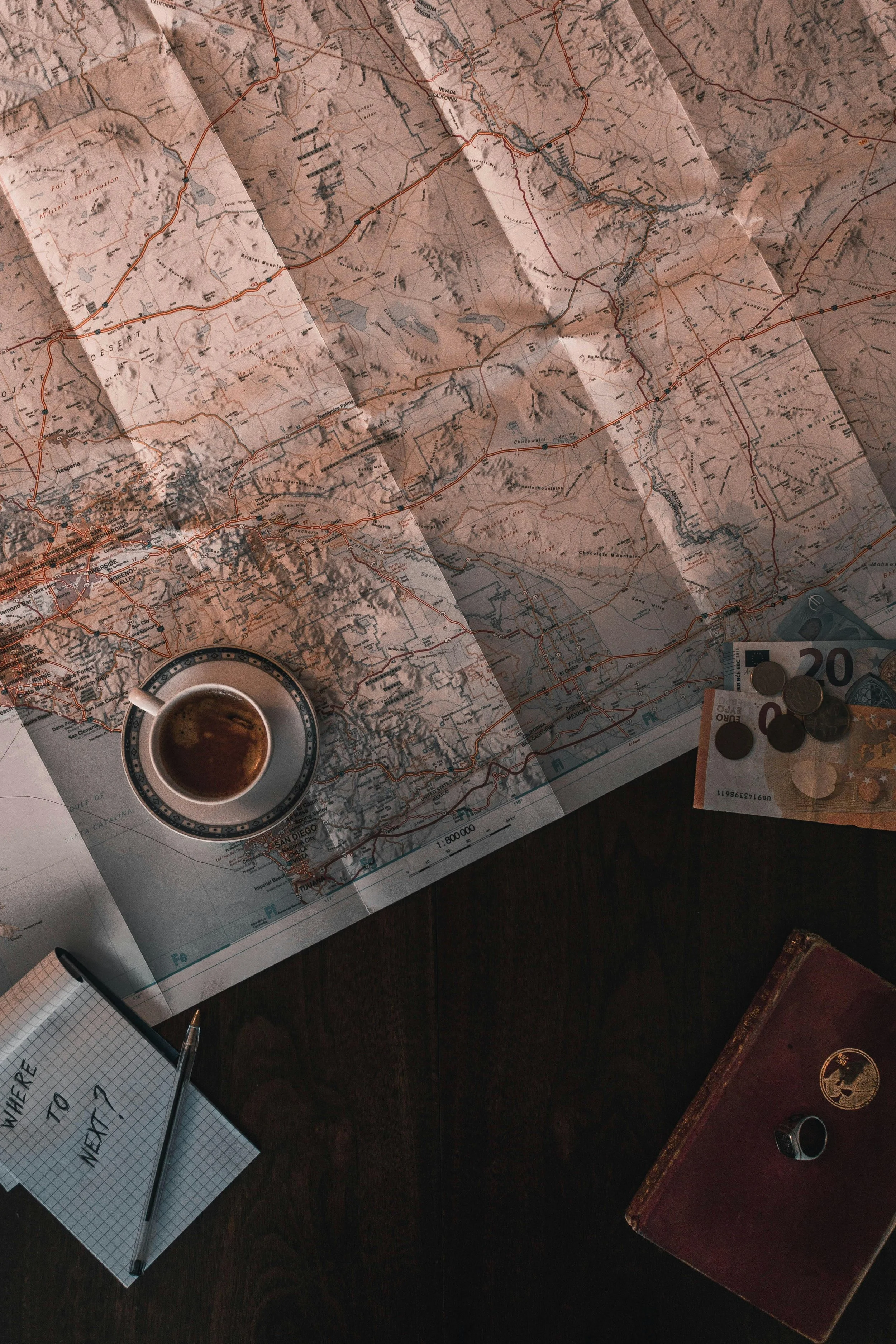 An overhead view of a travel planning scene with a map, a cup of coffee, currency notes, coins, a journal, and a notebook with a pen.
