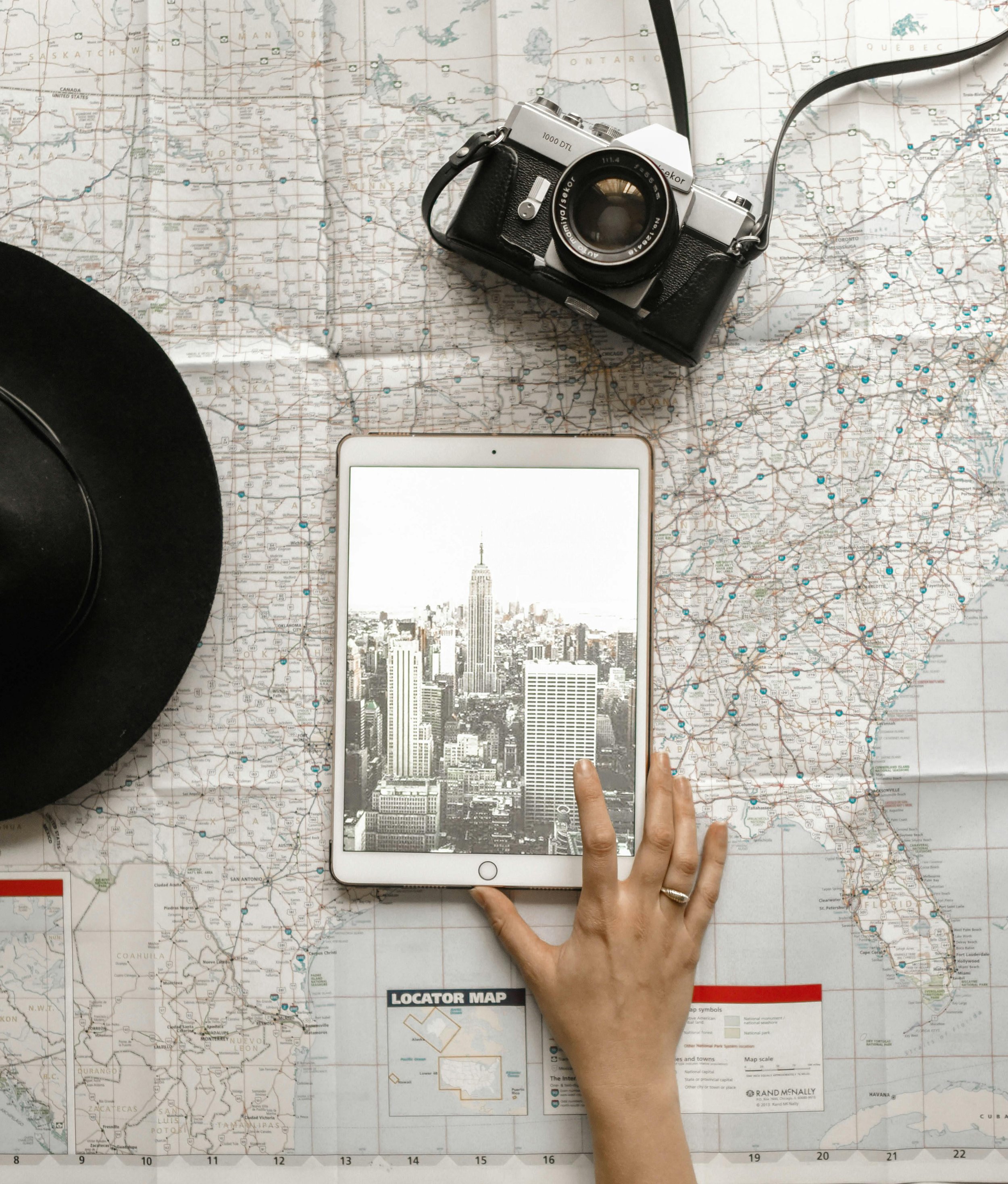 A map of the United States spread out on a table with a black hat, a vintage camera, a tablet displaying the Empire State Building, and a person's hand reaching towards the tablet.