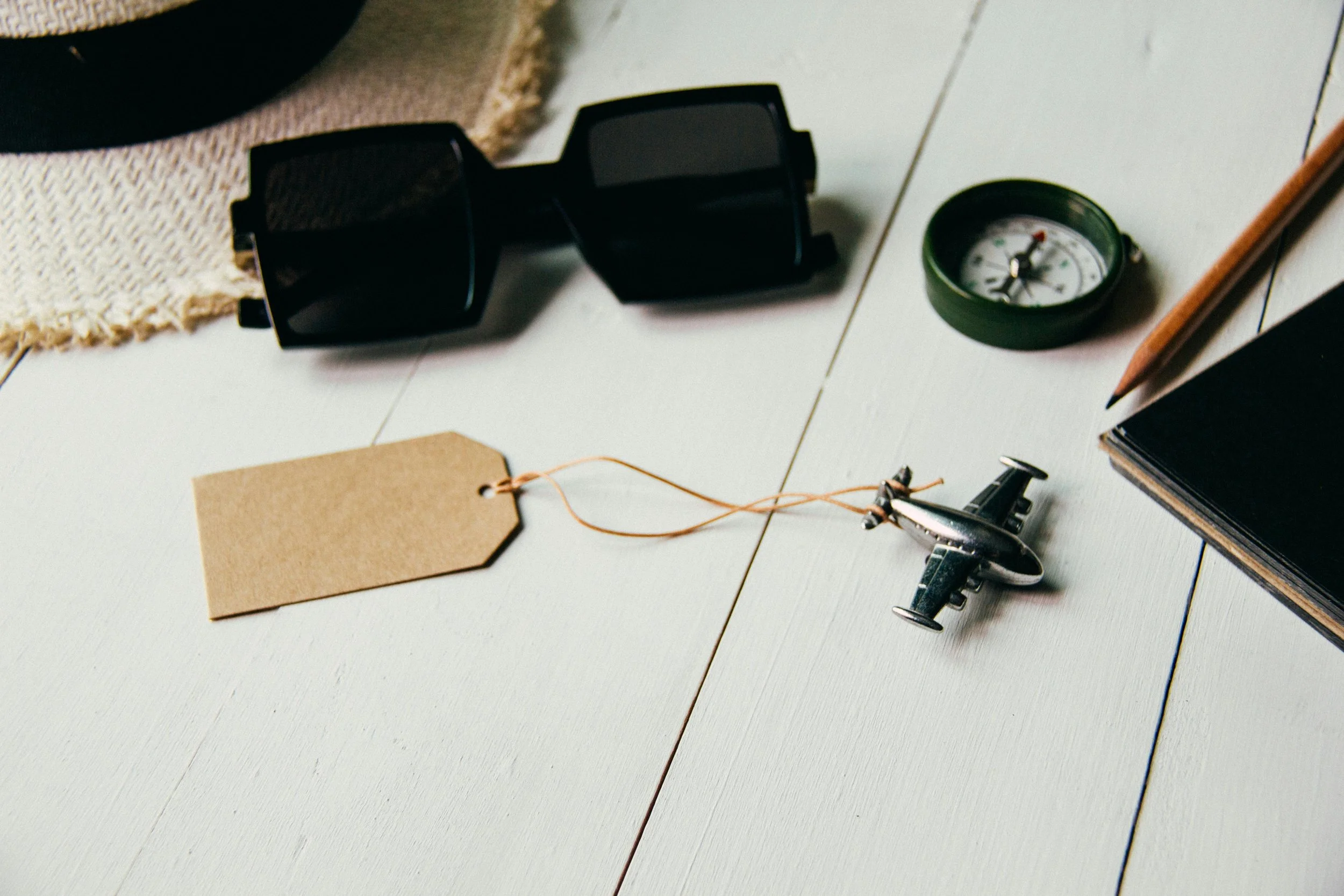 Collection of travel-related items on a white wooden surface including a pair of black sunglasses, a small compass, a vintage toy airplane, a blank brown luggage tag, a notebook, and a wooden pencil.