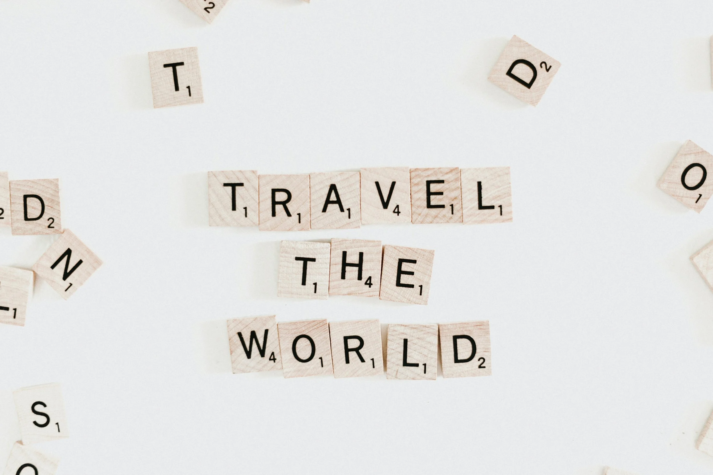 Scrabble tiles spell out 'Travel the World' on a white background with other scattered tiles around.