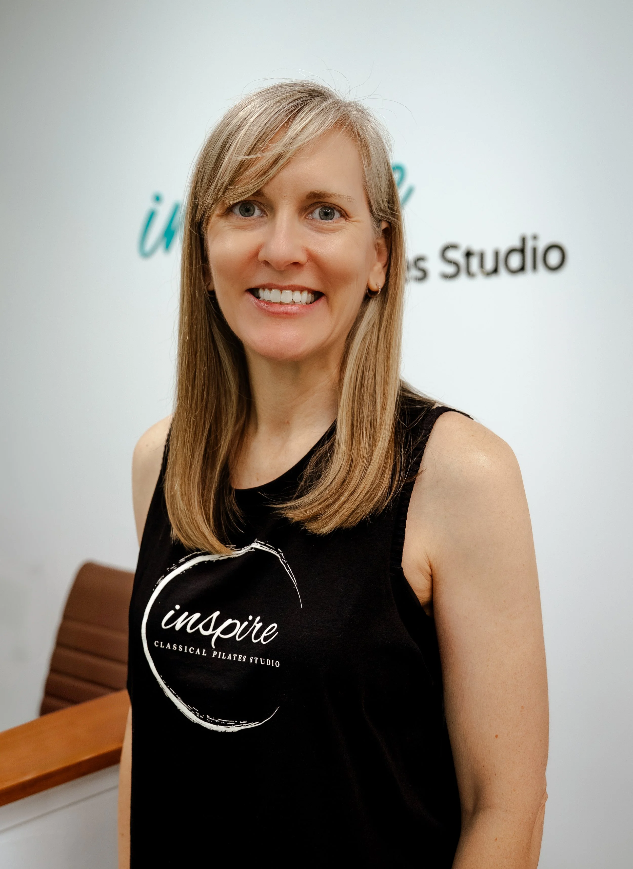 A woman with red hair styled in an updo, wearing a black sleeveless top, standing indoors in front of a beige wall with partially visible text. She is smiling and has one hand on her hip.