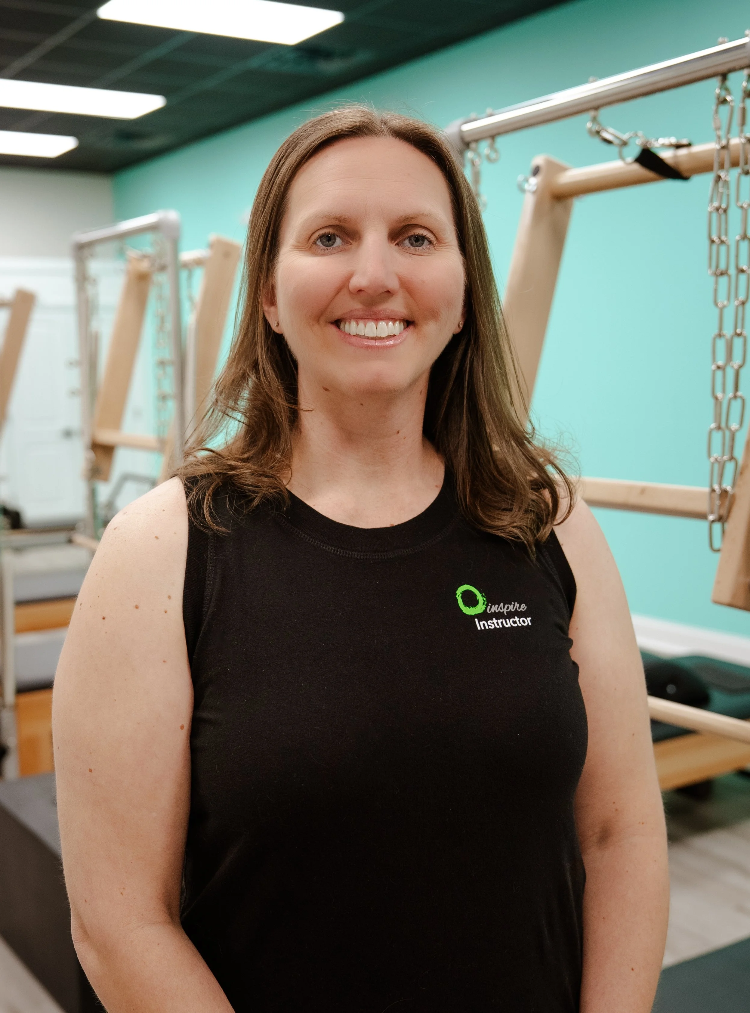 Portrait of a woman smiling with shoulder-length brown hair, wearing a light pink top, outdoors with green foliage in the background.