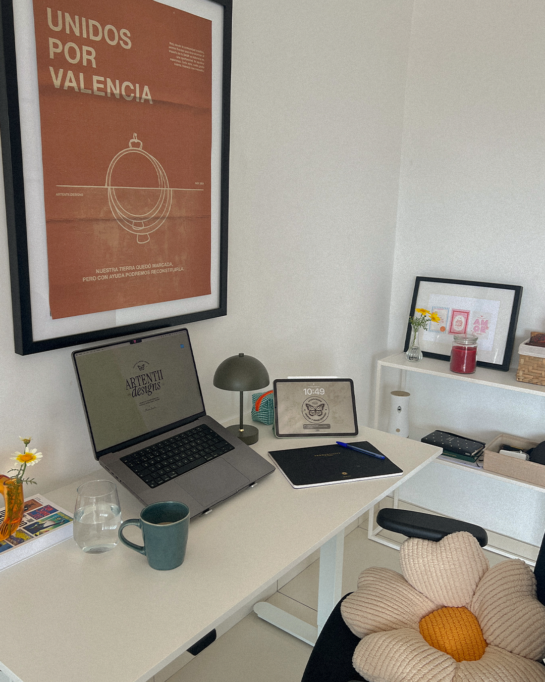 A home office desk setup with a laptop displaying a design website, a digital clock, a green table lamp, and a notebook with a pen. A mug and a glass of water are on the desk. A flower-shaped cushion is on the chair. There’s a framed poster on the wa