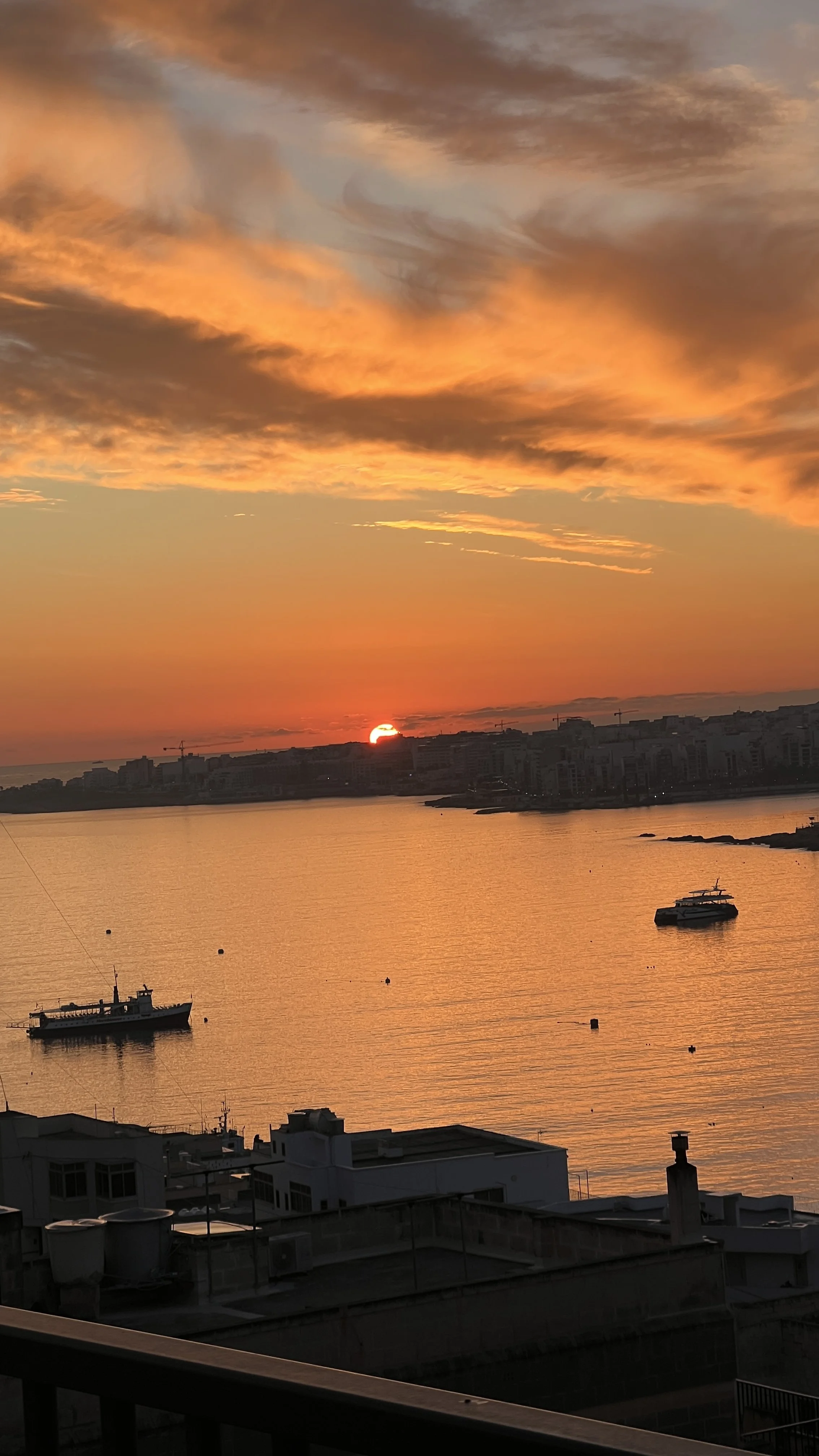 Scenic sunset over a calm bay with boats, silhouetted buildings, and dramatic clouds in the sky.