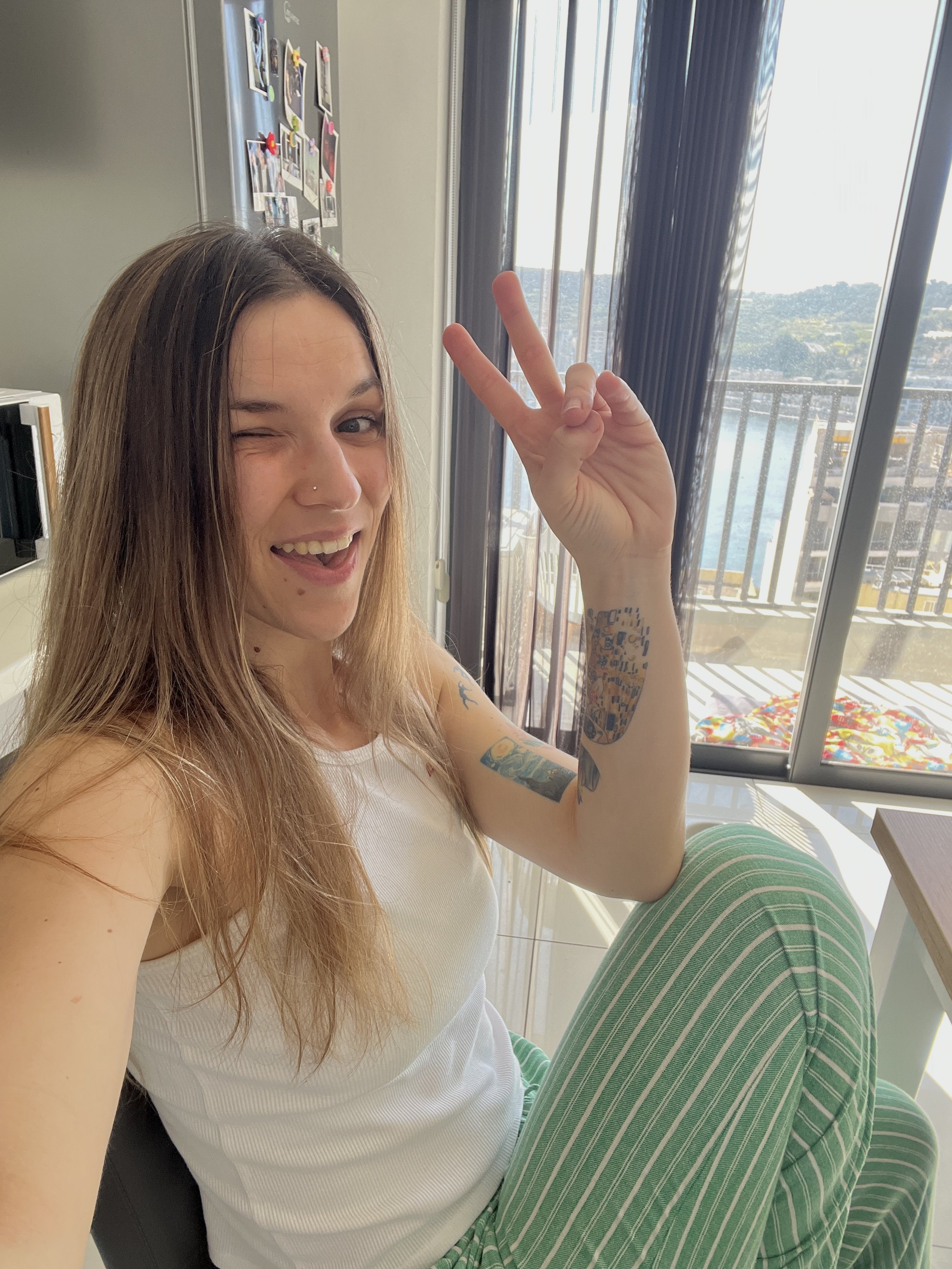 A woman with long hair winking and making a peace sign, wearing a white tank top and green striped pants, sitting indoors next to a window with a view of a balcony and scenery outside.