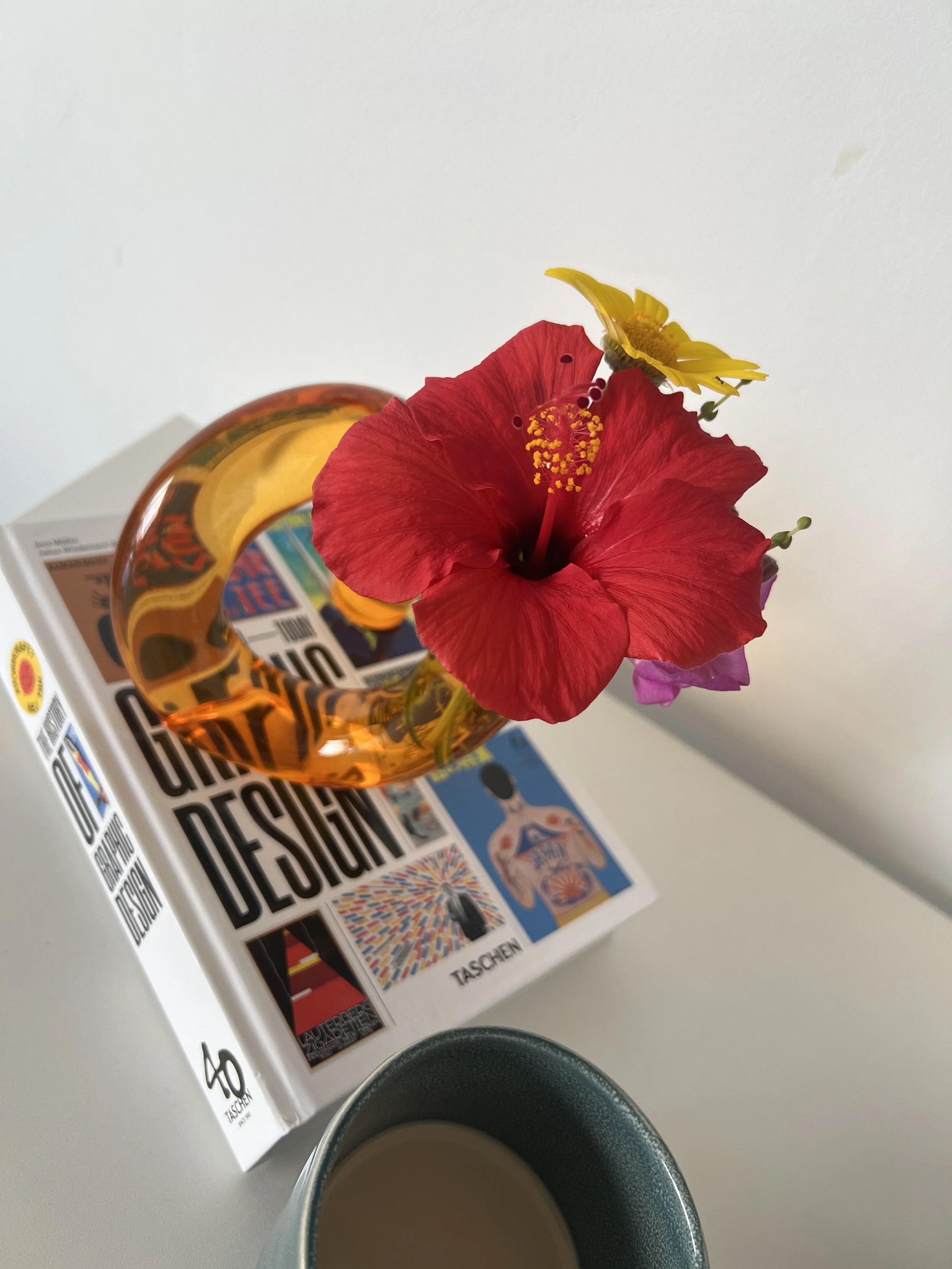 Red hibiscus and yellow flower in amber vase on book with coffee mug.
