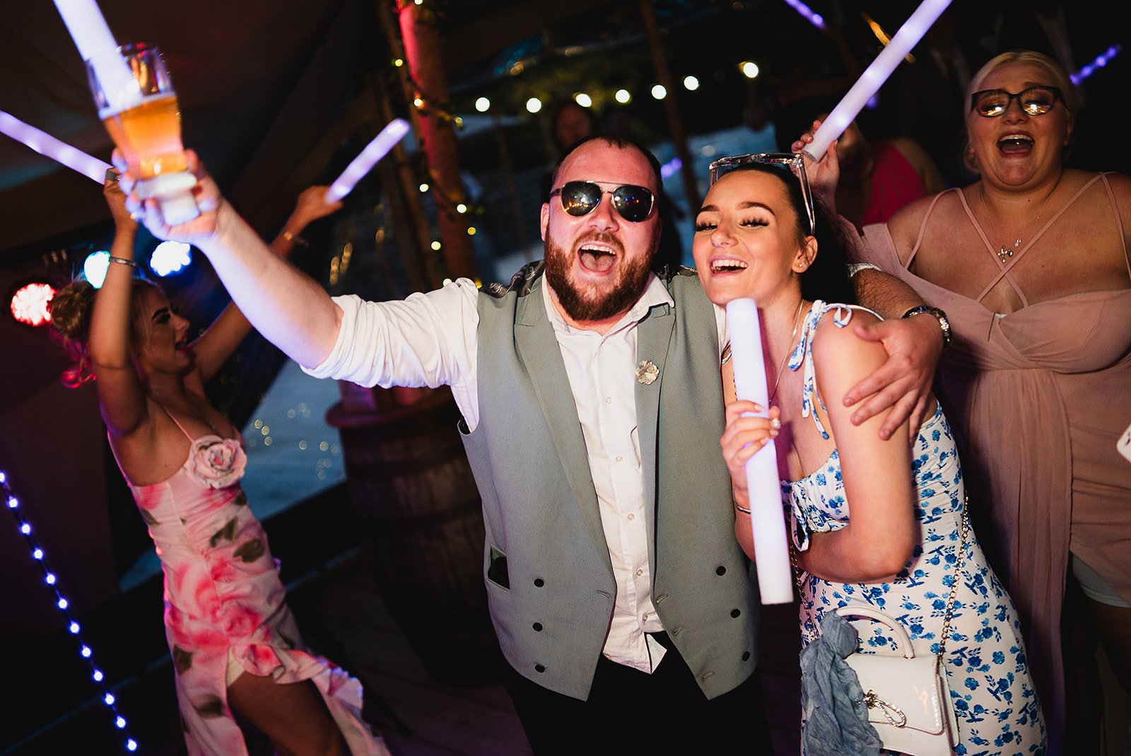 People enjoying a party, holding glow sticks and drinks, under colorful lights.