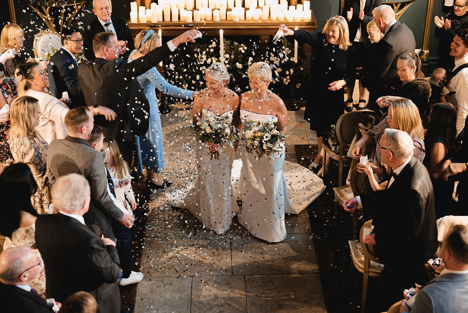 Wedding ceremony with confetti, two brides in white dresses holding bouquets, surrounded by smiling guests indoors, candles in background.