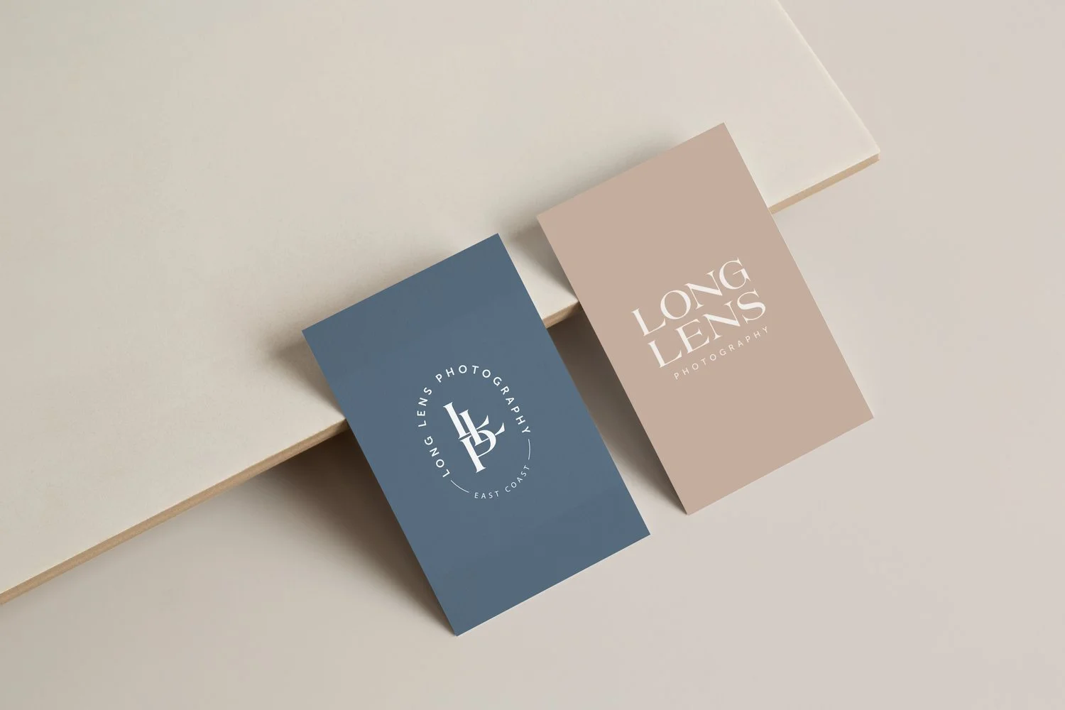 Two business cards on a light-colored surface, one dark blue and one beige, both with white text related to Long Lens Photography.