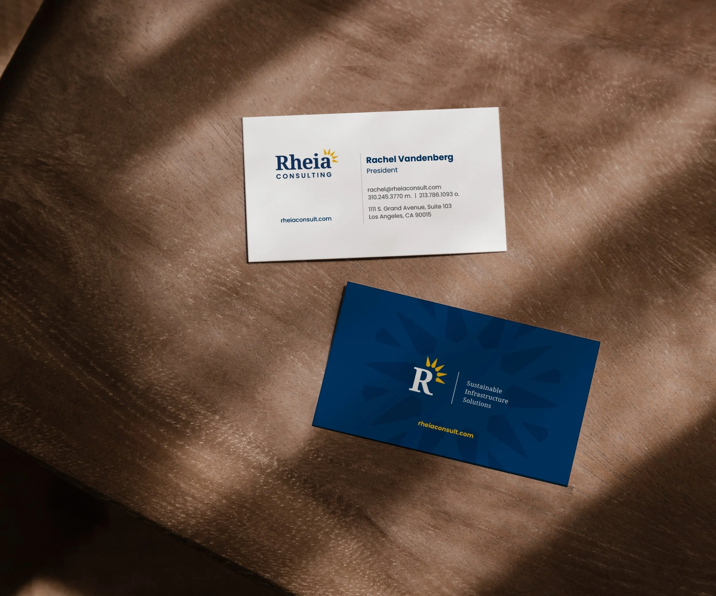 Business cards on a brown textured surface, one white with contact information for Rachel Vandenberg and one dark blue with the company's logo and website.