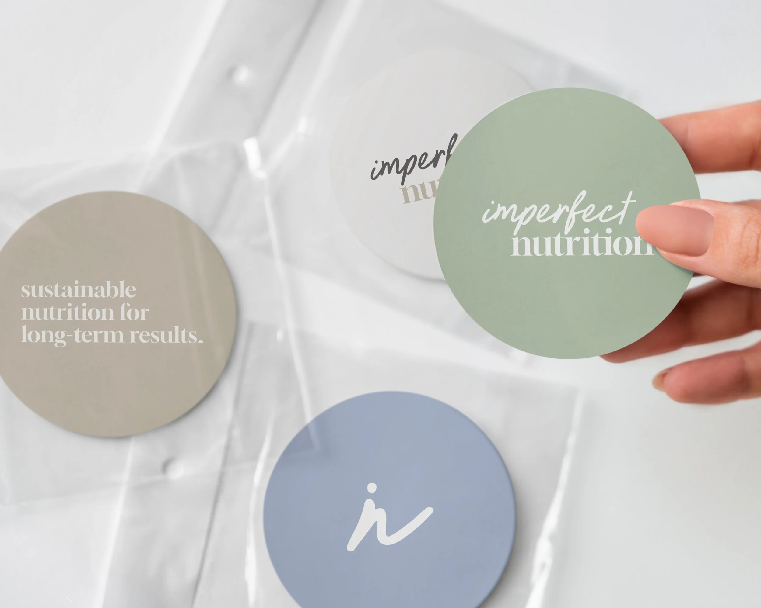 Person holding a green circular sticker with the words 'imperfect nutrition' on it, surrounded by other circular stickers with the phrases 'sustainable nutrition for long-term results' and 'imperfect human nutrition' on a white surface.