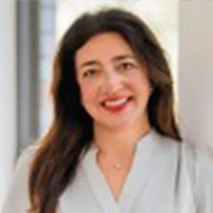 A woman with long dark hair smiling, wearing a light-colored blouse, standing indoors near a window.