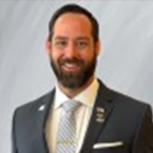A man with a beard and mustache wearing a dark suit, light-colored shirt, and patterned tie, standing against a light background.