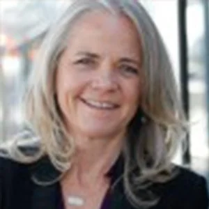Portrait of a smiling older woman with shoulder-length gray hair, wearing a dark top, outdoors with a blurred background.