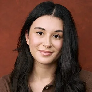 Portrait of a young woman with long dark hair and a subtle smile, wearing a brown top, against a reddish-brown background.