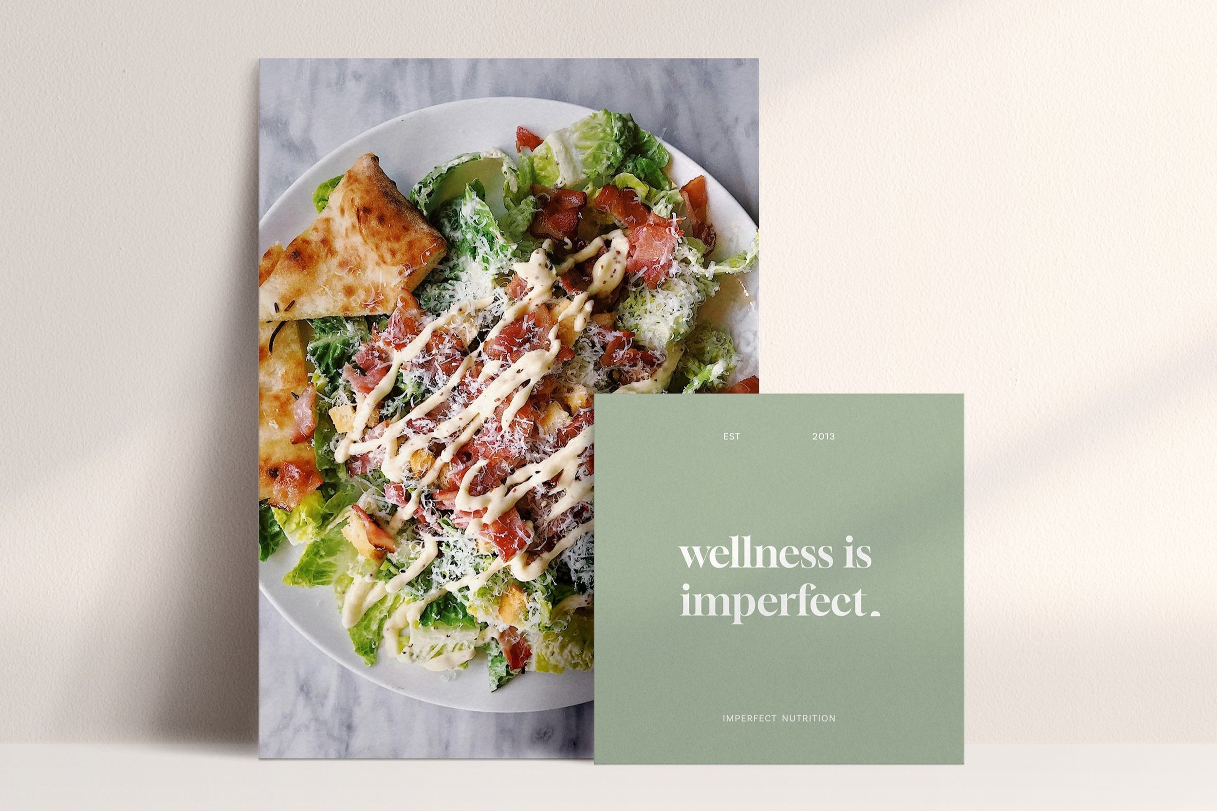 A printed card with the phrase 'wellness is imperfect' partially covers a photo of a salad with lettuce, tomatoes, shredded cheese, dressing, and a slice of bread, all on a white plate.