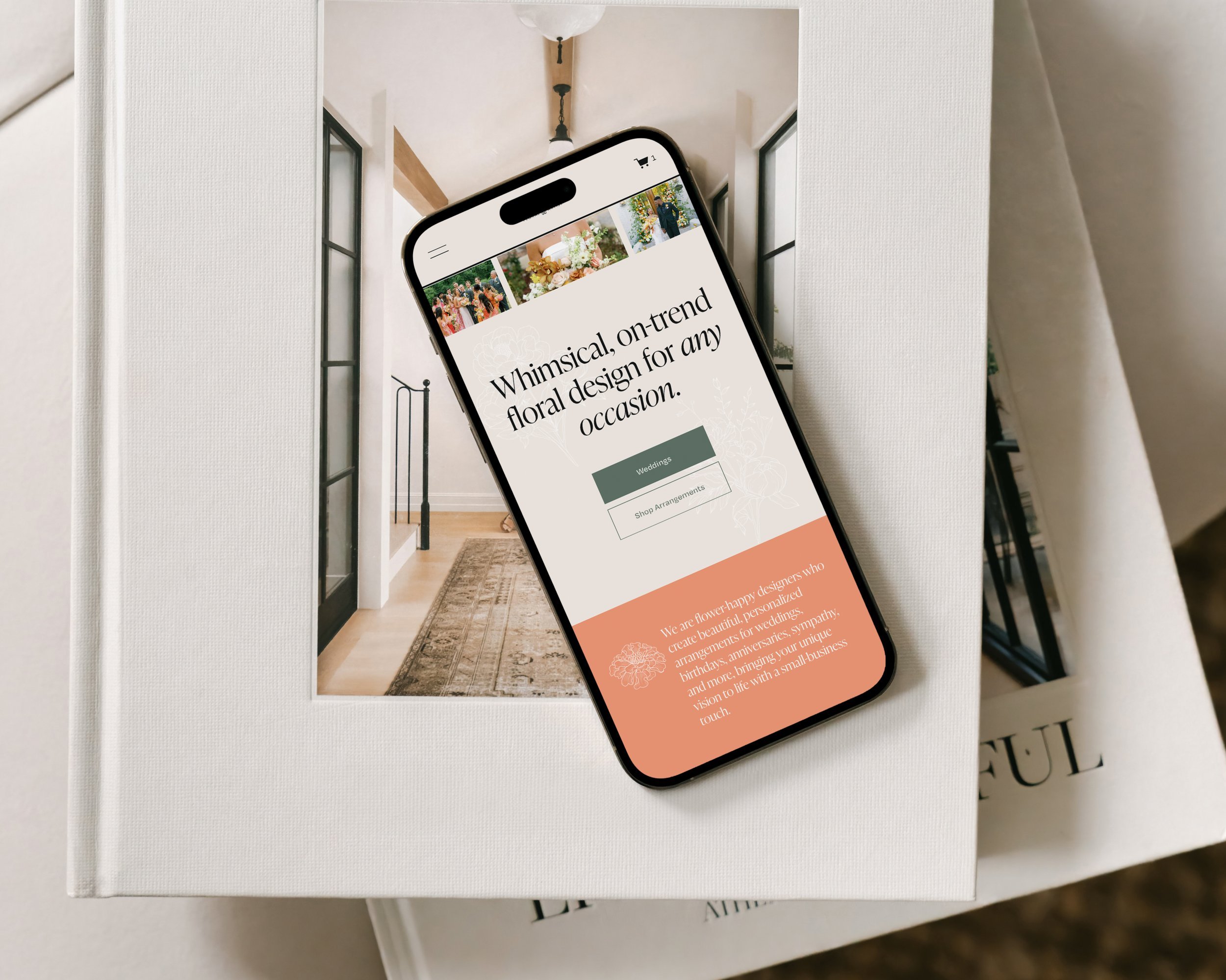 A smartphone displaying a floral design website, resting on a white book with a partial view of a second book underneath, on a light-colored table.