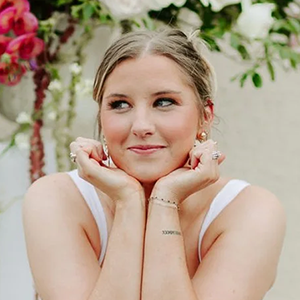 A young woman with blonde hair and a playful expression, resting her chin on her hands, standing outdoors with flowers and greenery in the background.